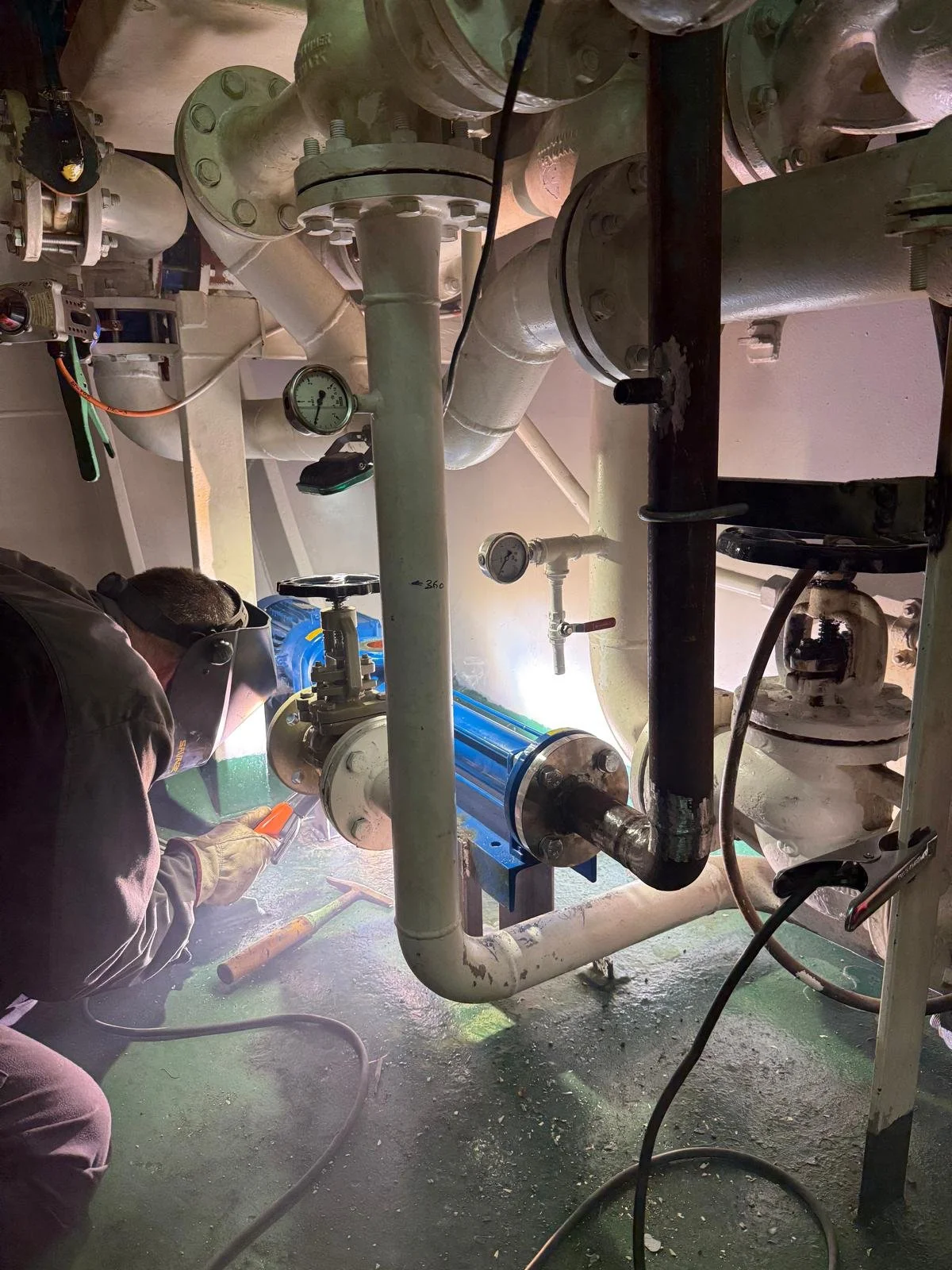 A worker welding a pipe in an industrial setting with various pipes and gauges around.
