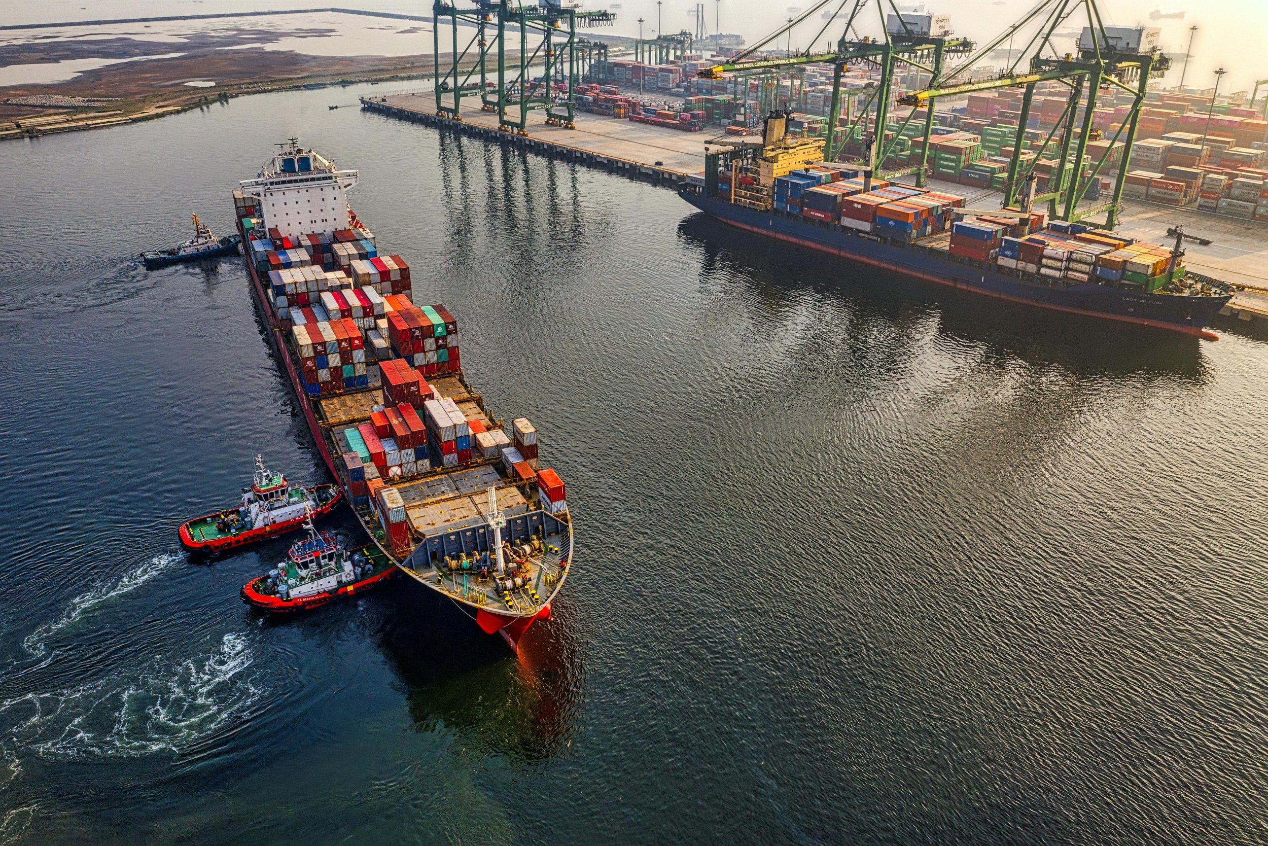 An aerial view of a busy shipping port with large cargo ships carrying containers, cranes loading and unloading containers, and tugboats guiding a ship through the water at sunset.
