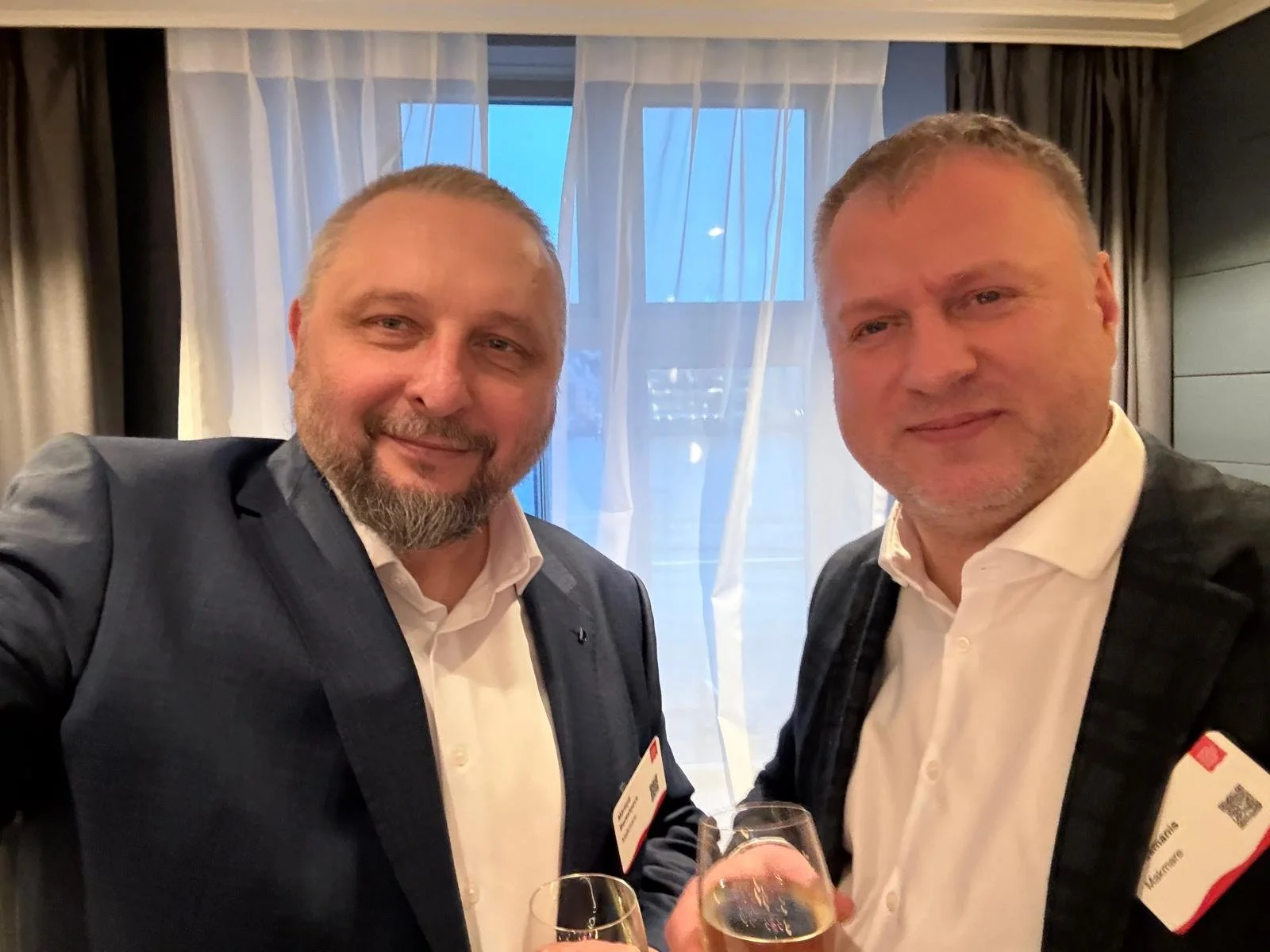 Two middle-aged men in suits holding glasses of champagne, smiling at the camera, standing in front of a window with sheer curtains at what appears to be a formal event.