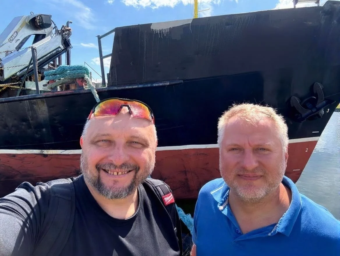 Two men taking a selfie in front of a large black and red boat docked on the water, smiling under a bright blue sky.