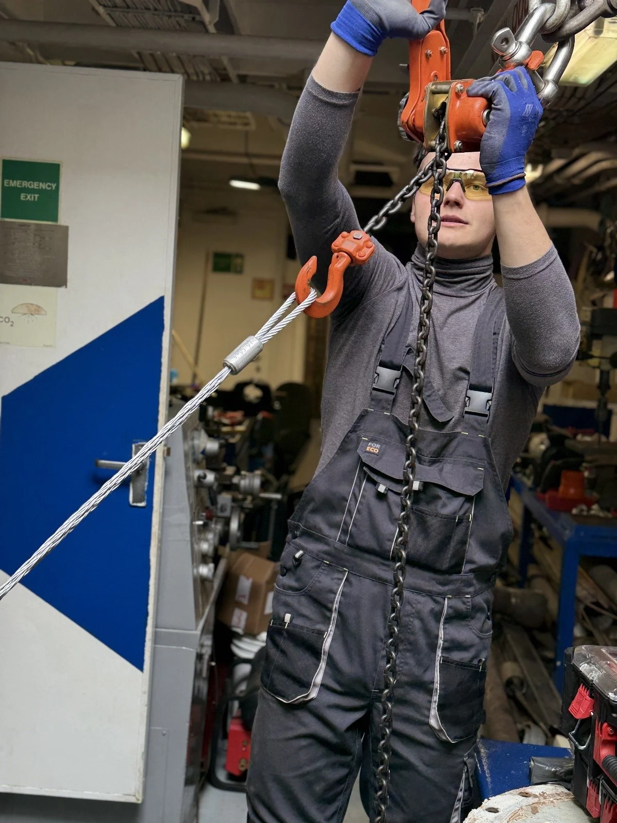 Person working in a workshop, using safety gloves and goggles, operating heavy-duty equipment with chains, shelves with tools and machinery in the background.