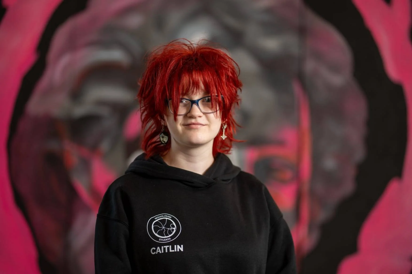 A woman with bright red, messy hair, wearing glasses, earrings, and a black hoodie with the name 'Caitlin' on it, standing in front of a large, colorful portrait of a person's face with a pink background.