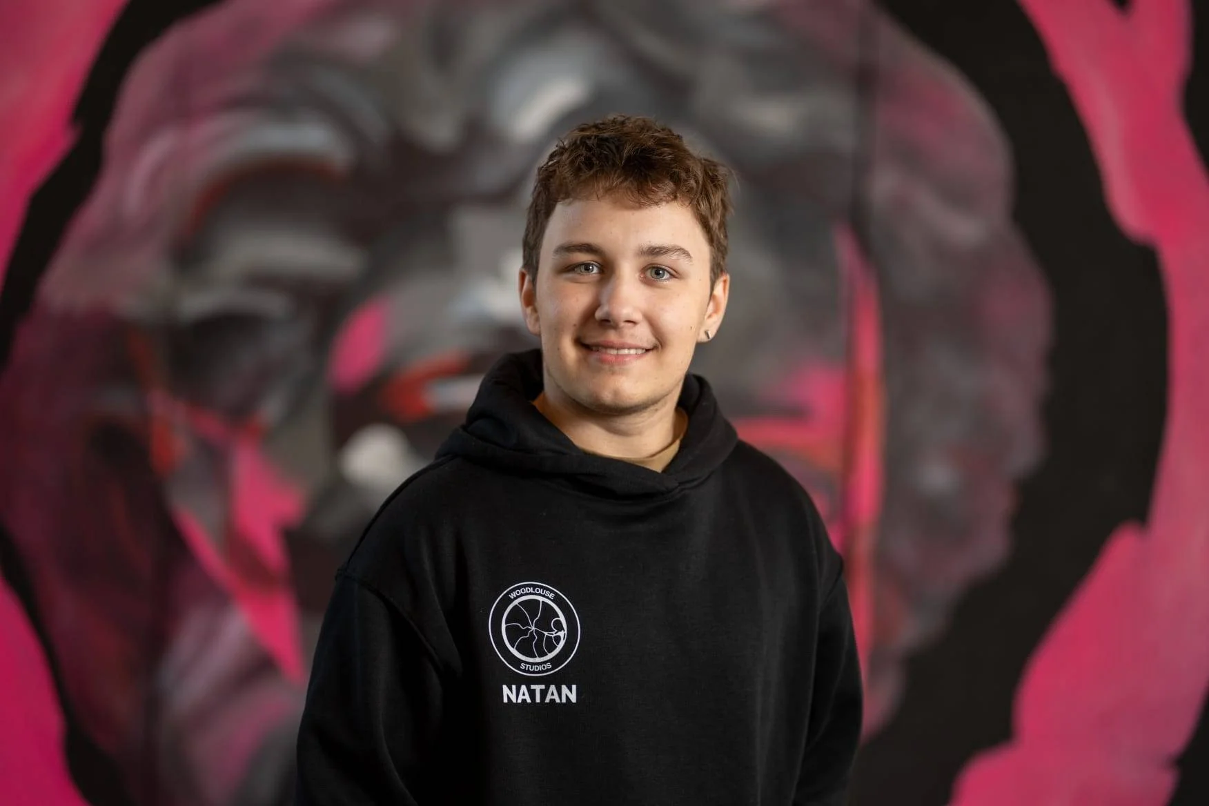 A young man with short brown hair and a black hoodie featuring a logo that reads 'WOODLOUSE STUDIOS' and the name 'NATAN' below. He is smiling and standing in front of a colorful mural of a human brain with pink and black accents.