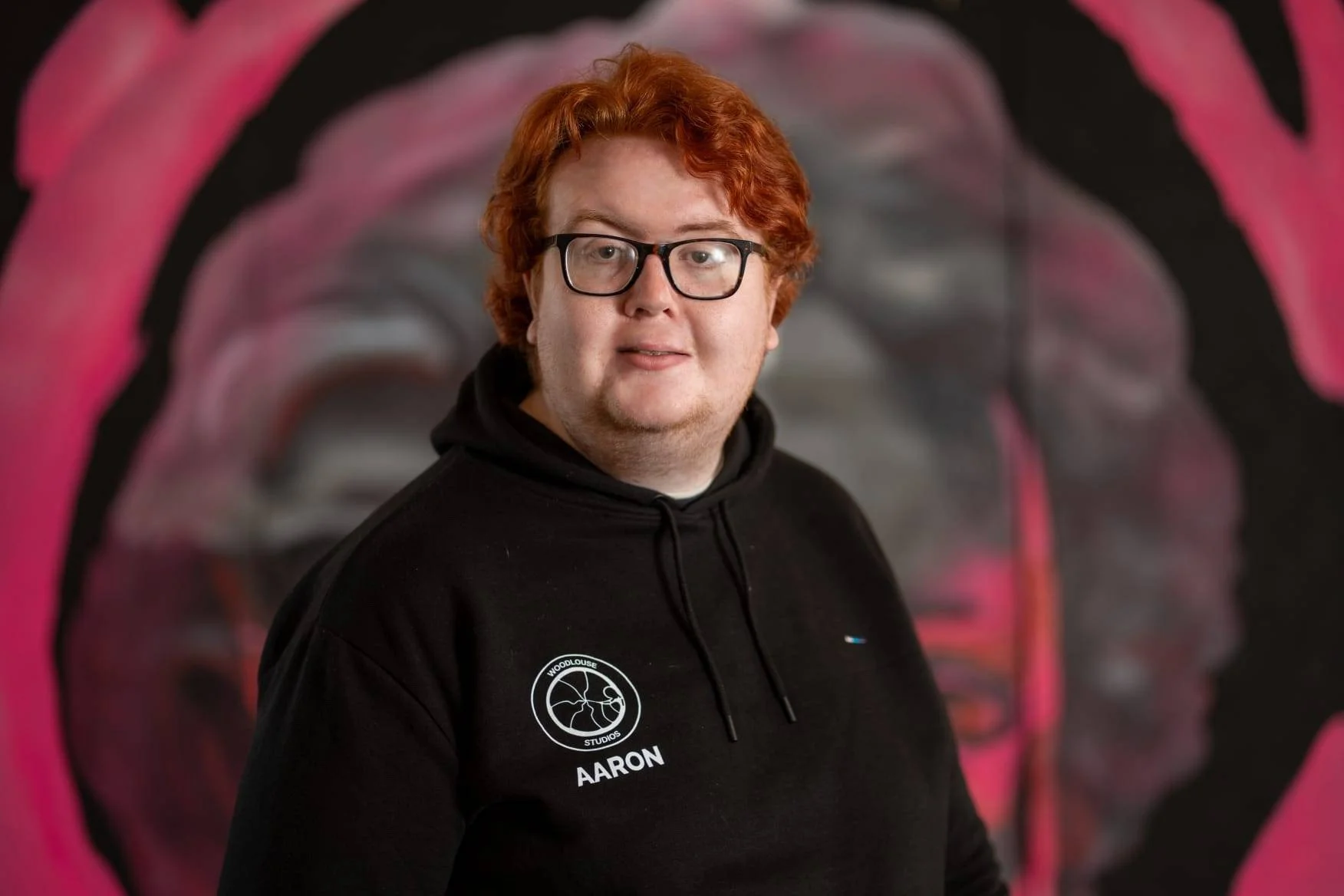 A man with red hair and glasses wearing a black hoodie with a logo and the name 'Aaron' on it, standing in front of a colorful abstract mural.