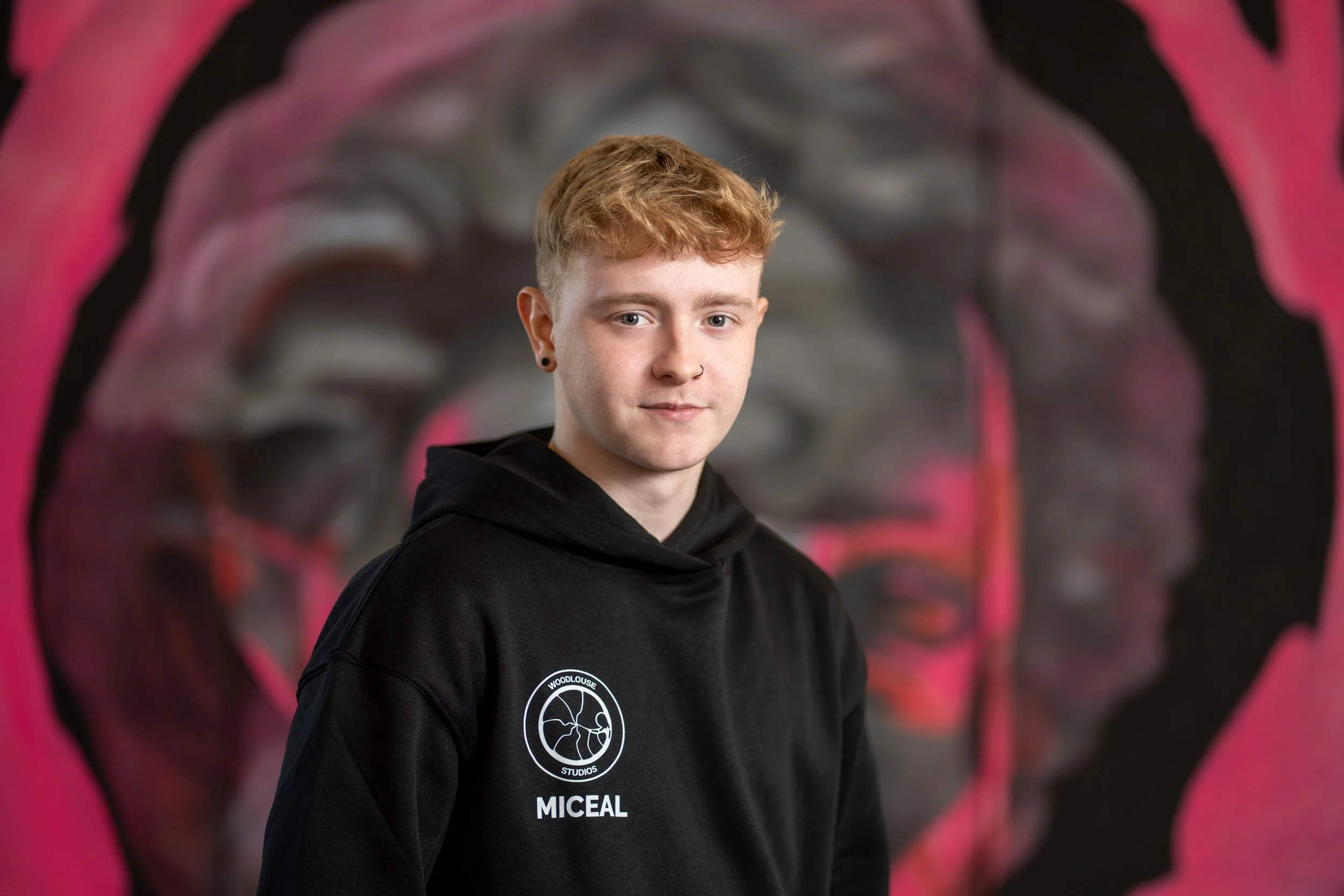 Young man with short red hair wearing a black hoodie with a logo and the name 'MICEAL' standing in front of a mural of a brain with pink, black, and gray colors.