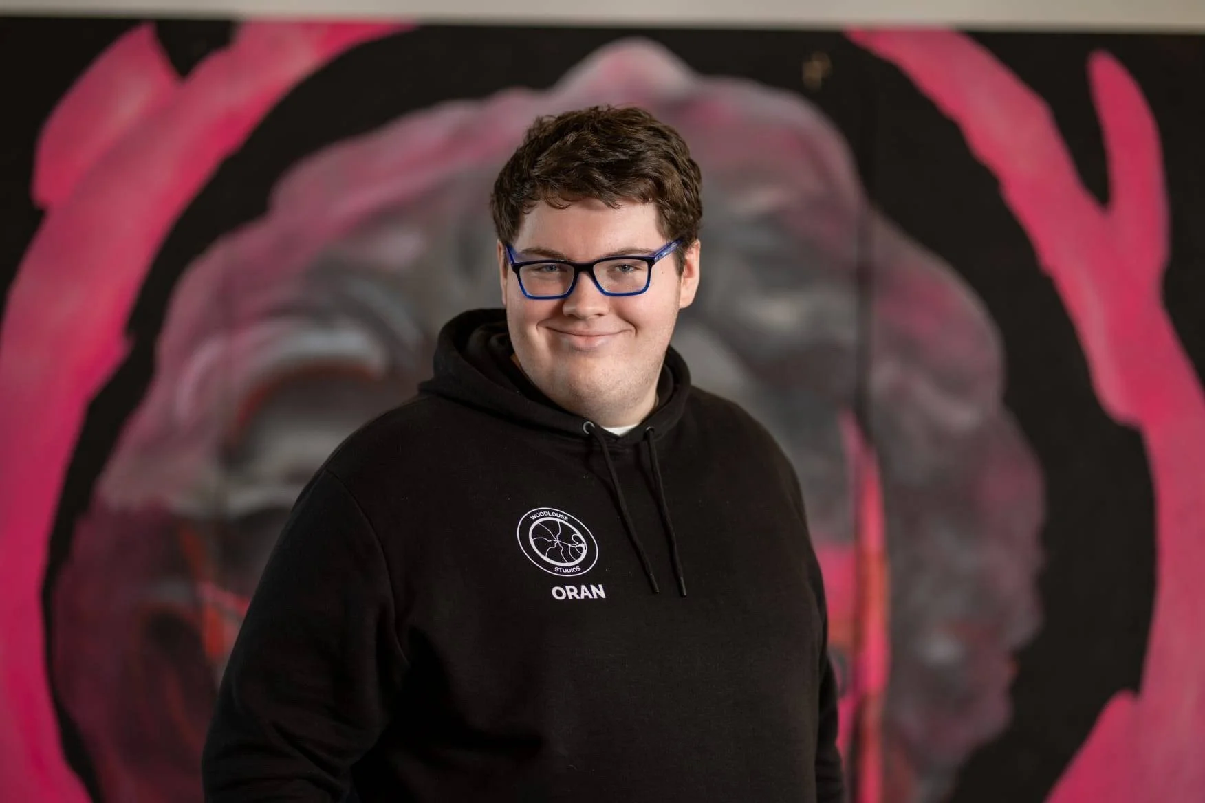 A young man with glasses and short brown hair, wearing a black hoodie with a logo and the name 'Oran' on it, standing in front of a pink and black graffiti-style mural.