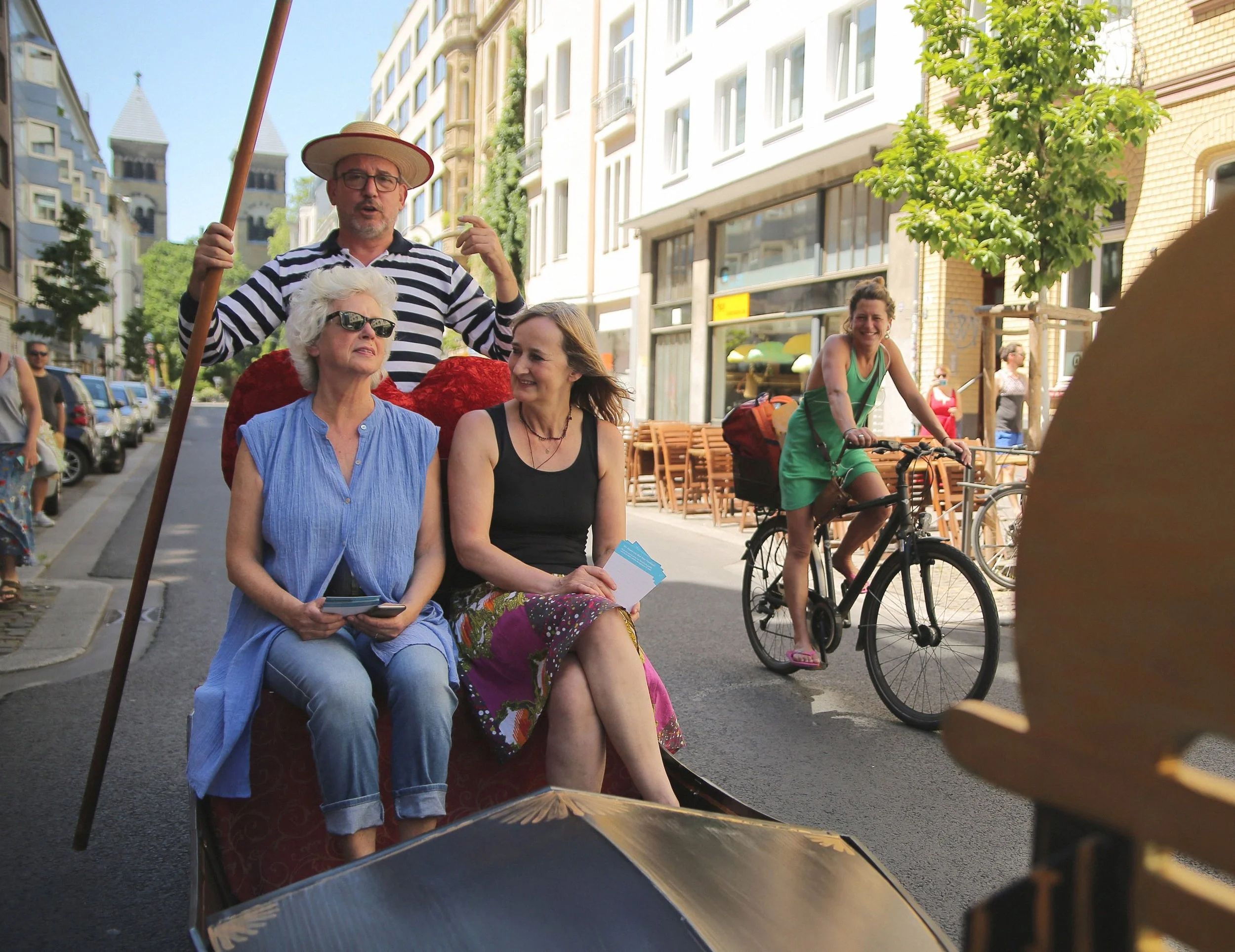 Eine Gruppe von fünf Personen erlebt eine Straßenparade. Zwei Frauen sitzen auf einem Kutschenwagen, eine trägt Sonnenbrille und hält ein Smartphone. Ein Mann steht hinter ihnen und spricht, während eine junge Frau auf einem Fahrrad vorbeifährt und in die Kamera lächelt.