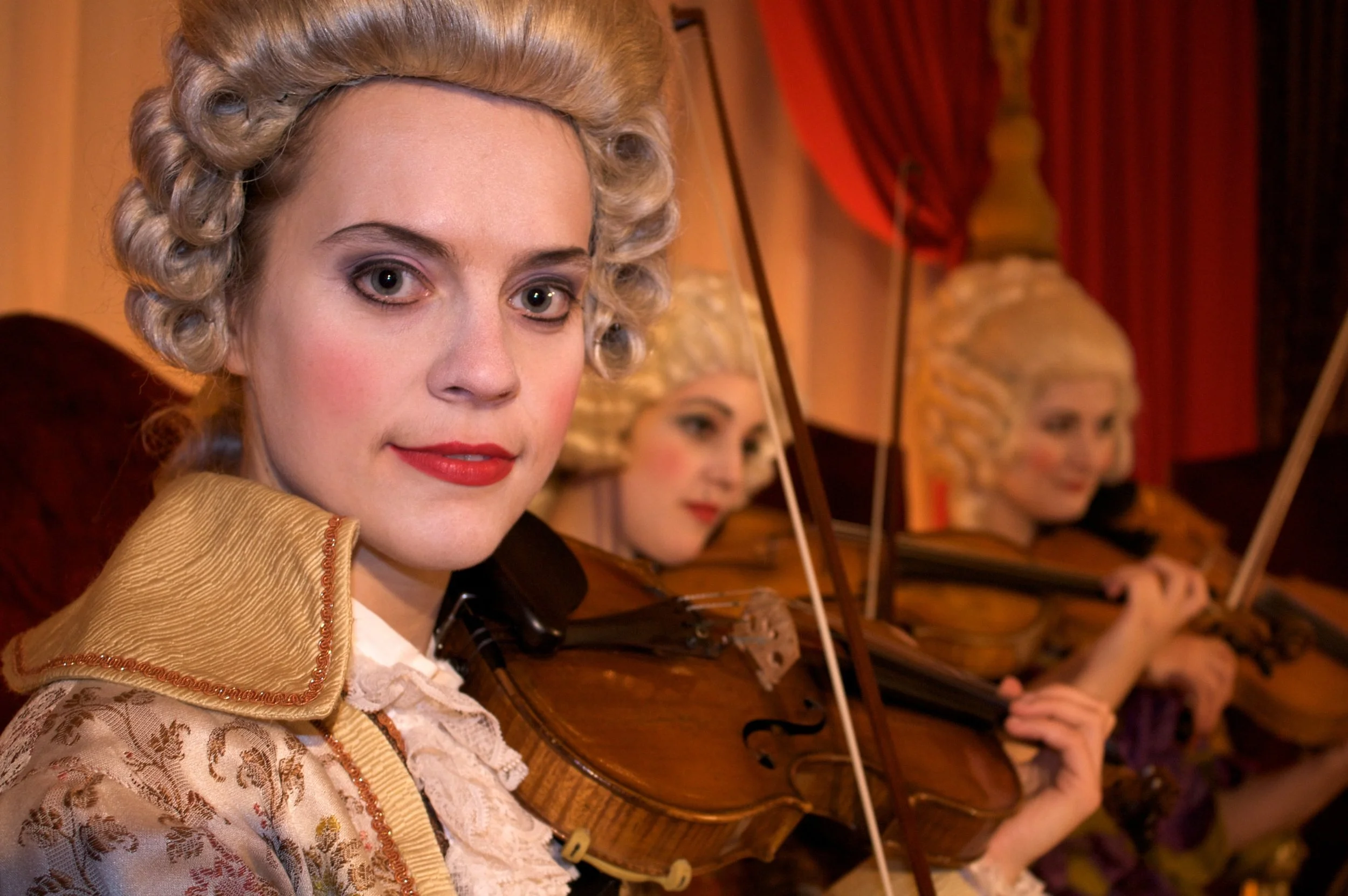Drei Frauen in historischer Kleidung spielen Streichinstrumente in einem Orchestersaal, im Vordergrund eine Frau mit Platinblonden Locken und rotem Lippenstift, im Hintergrund zwei weitere Frauen.