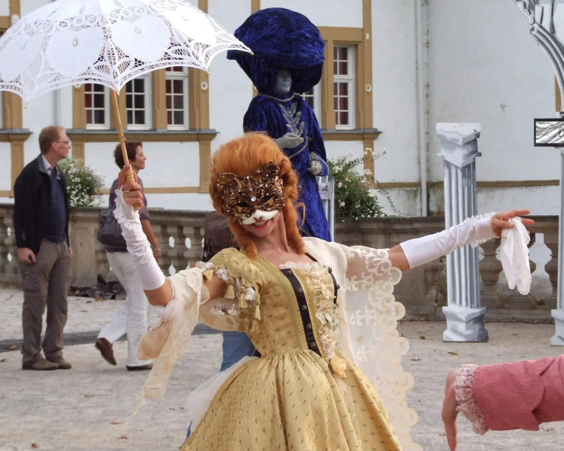 Eine Frau in einem historischen gelben Kleid mit einer floralen Maske und einer Perücke posiert bei einem Fest. Sie hält einen weißen doppelten Regenschirm in der rechten Hand und eine weiße Stoffbahn in der linken Hand. Im Hintergrund sind andere Personen, ein Gebäude mit Fenster und eine Skulptur zu sehen.