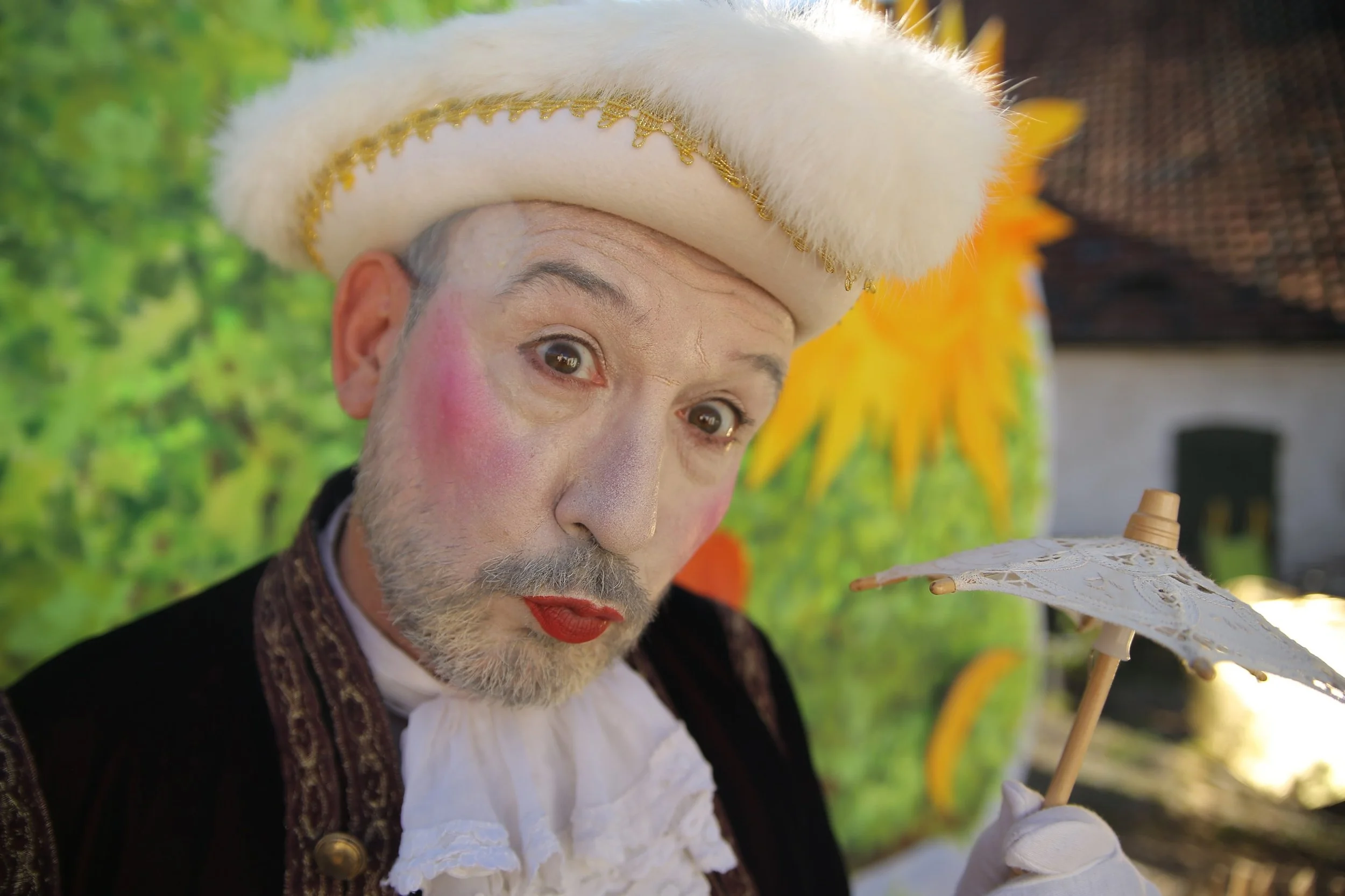 Ein Mann im elaborierten Kostüm mit bunter Schminke und Hut, der ein kleiner Sonnenschirm hält, bei einer festlichen Veranstaltung im Freien.