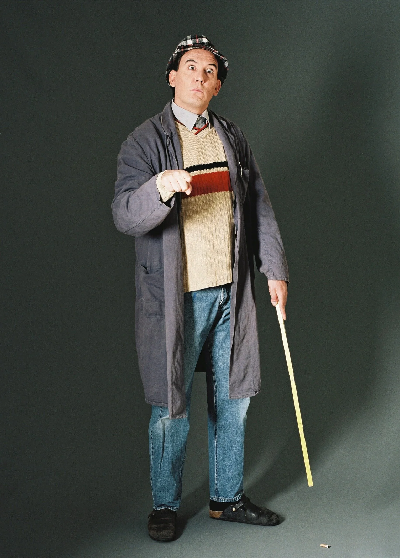 Ein Mann in Kleidung im Stil eines Charakters aus einem alten Film, trägt eine Mütze, eine Jacke, eine Pullover und Jeans, hält einen Meterstab und einen Zigarettenstummel, steht vor einem dunklen Hintergrund.