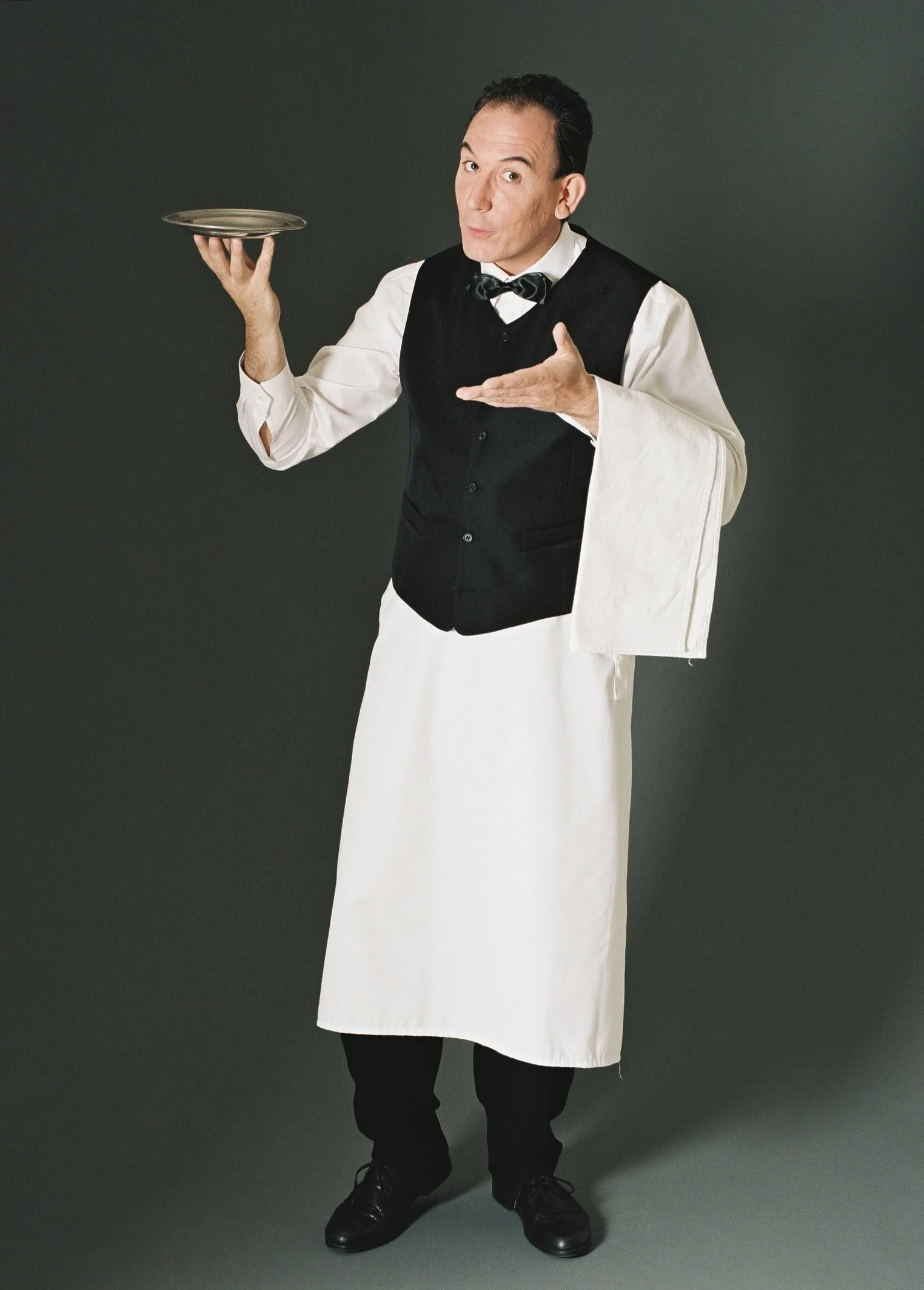 Mann in Kellneruniform hält ein silbernes Teller und zeigt eine Einladung mit einer Hand, vor dunklem Hintergrund.