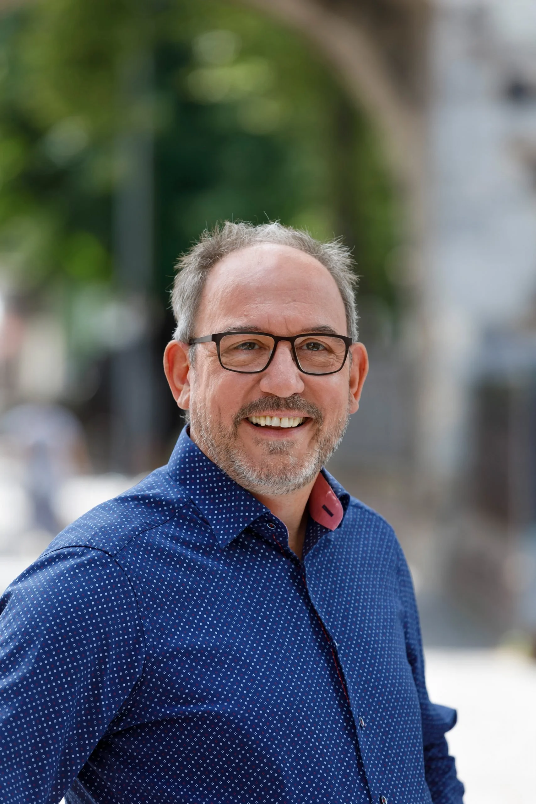 Ein lachender Mann mit Brille, Kurzhaar, barfuß, trägt eine blaue Hemd mit kleinen weißen Punkten, steht vor einem urbanen Hintergrund.
