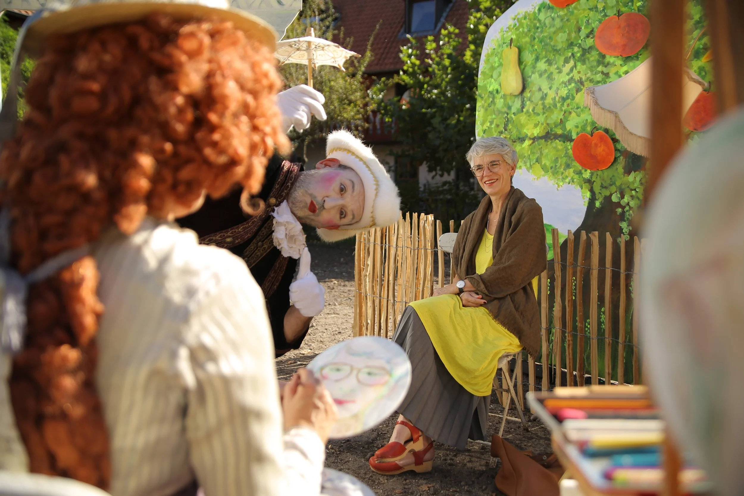 Eine Frau sitzt lachend vor einem bunten Wandbild eines Obstbaums. Zwei verkleidete Personen, eine mit Clownskostüm und die andere mit gelber Kleidung und brauner Jacke, posieren vor ihr. Es sieht nach einer Outdoor-Veranstaltung aus.