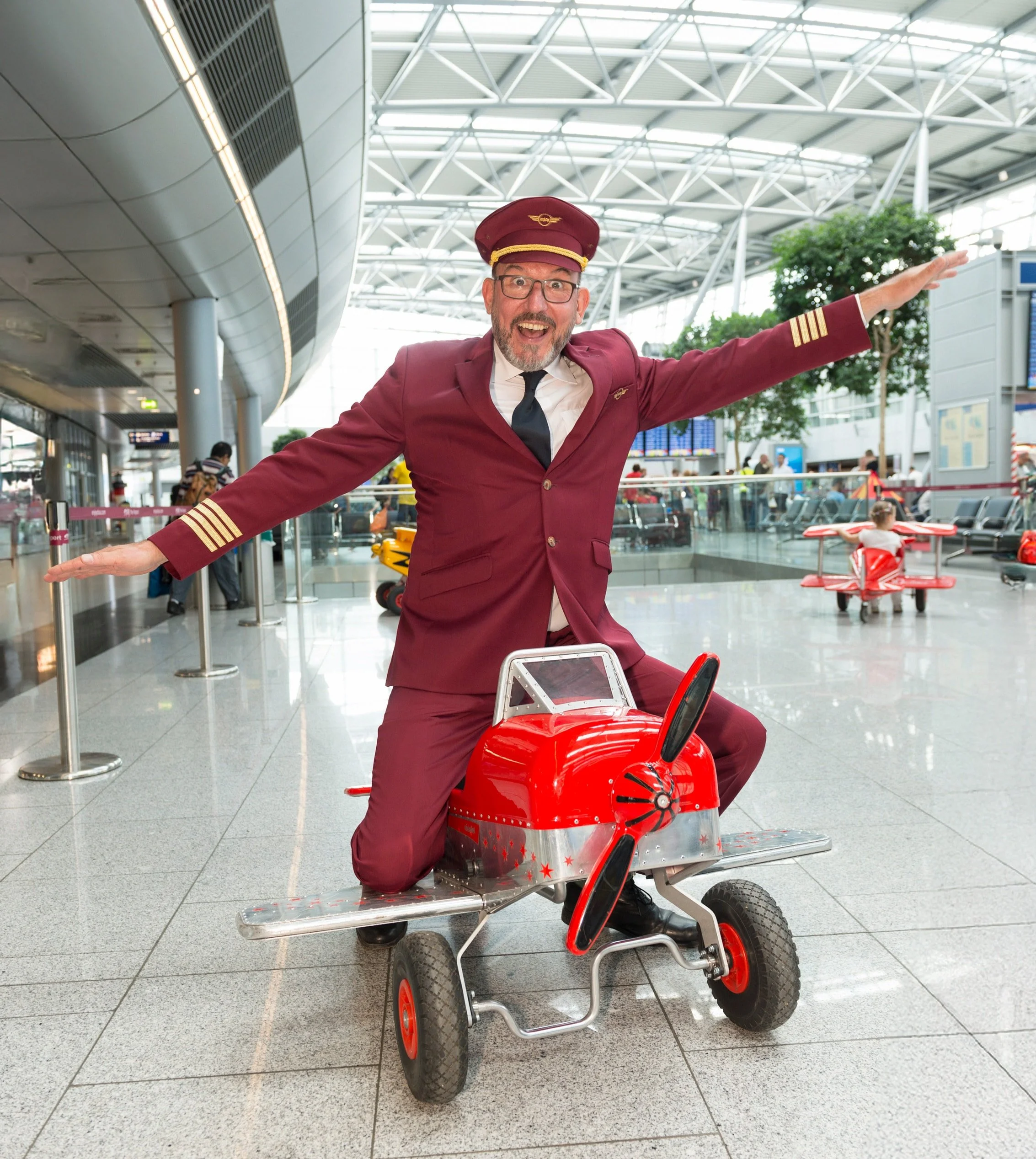 Mann im Flugbegleiter-Outfit auf einem Kinderfahrzeug in einem Flughafen