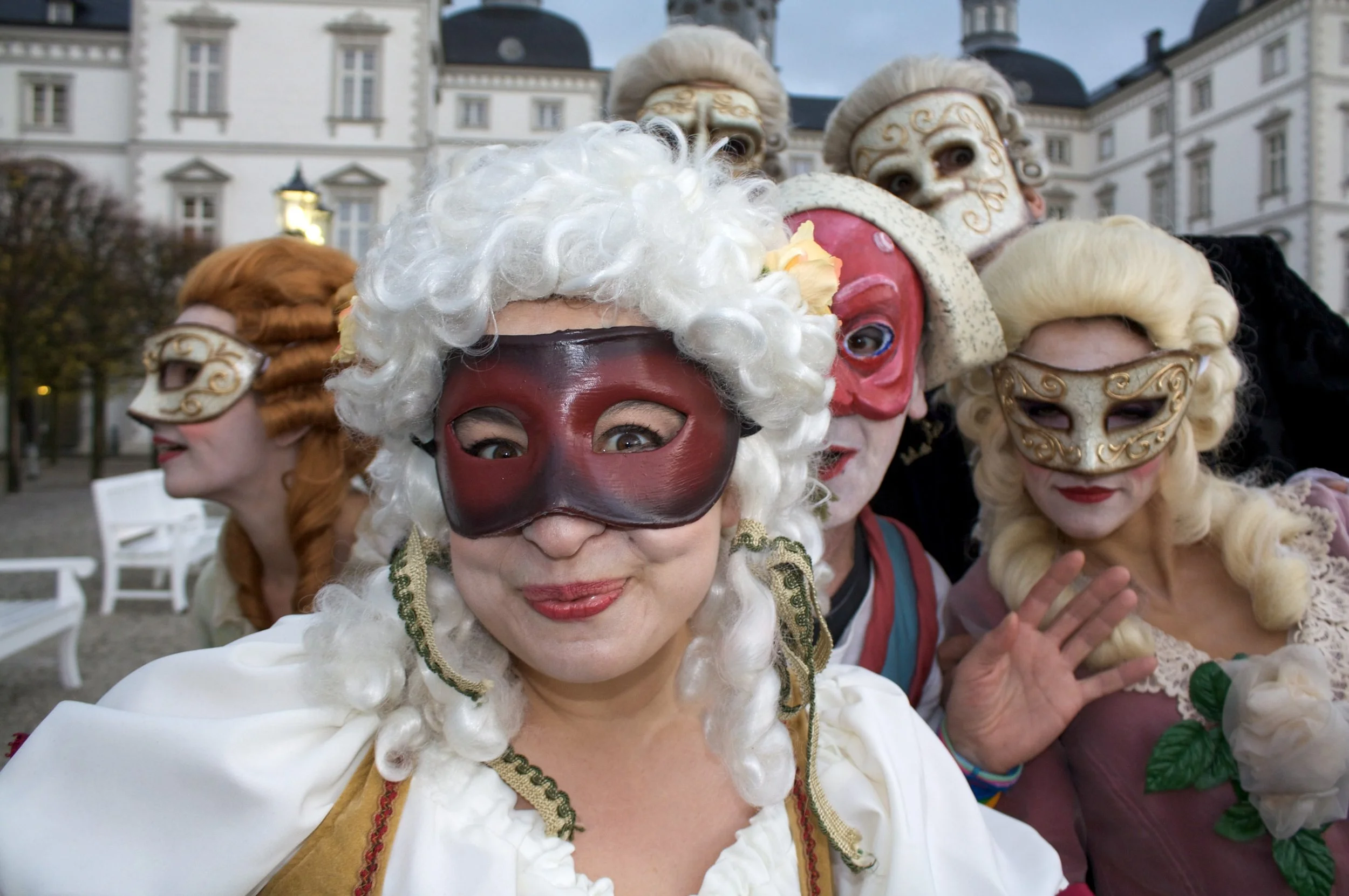 Menschen in farbenfrohen Kostümen und Masken bei einer Karnevalsveranstaltung im Freien, im Hintergrund ein historisches Gebäude.