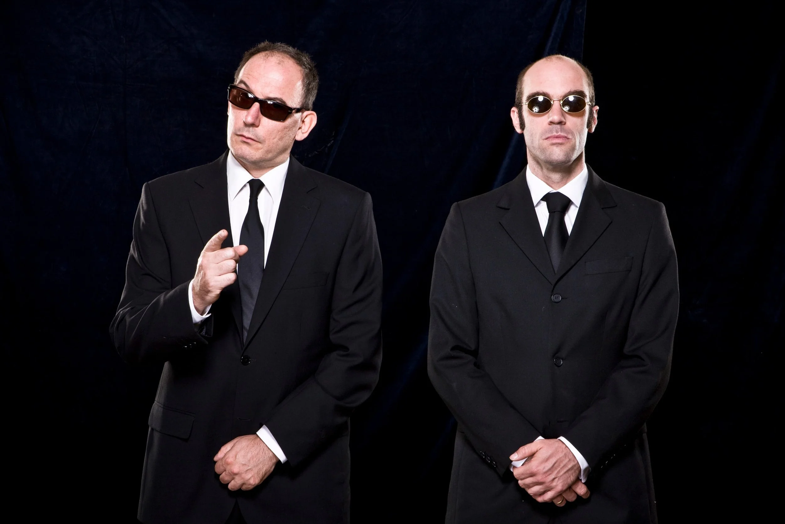 Zwei Männer in schwarzen Anzügen und Krawatten, beide tragen Sonnenbrillen, stehen vor schwarzem Hintergrund, einer zeigt auf die Kamera, der andere hat die Hände gefaltet.