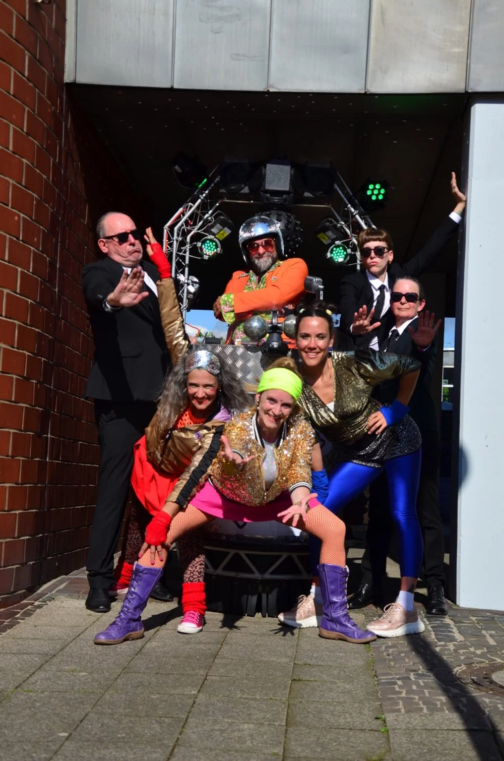 Gruppe von Menschen in bunten, auffälligen Kostümen posiert vor einem DJ-Setup im Freien.