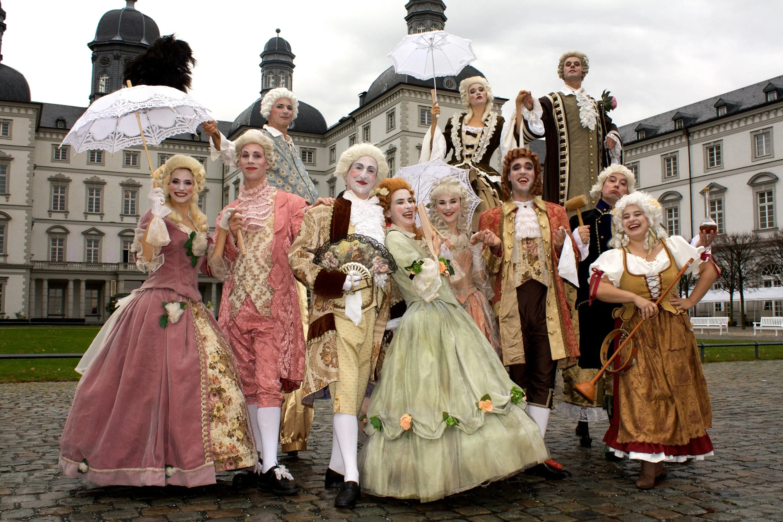 Gruppierung von Menschen in Barockkostümen, die an einem historischen Ort mit großen Gebäuden im Barockstil stehen.