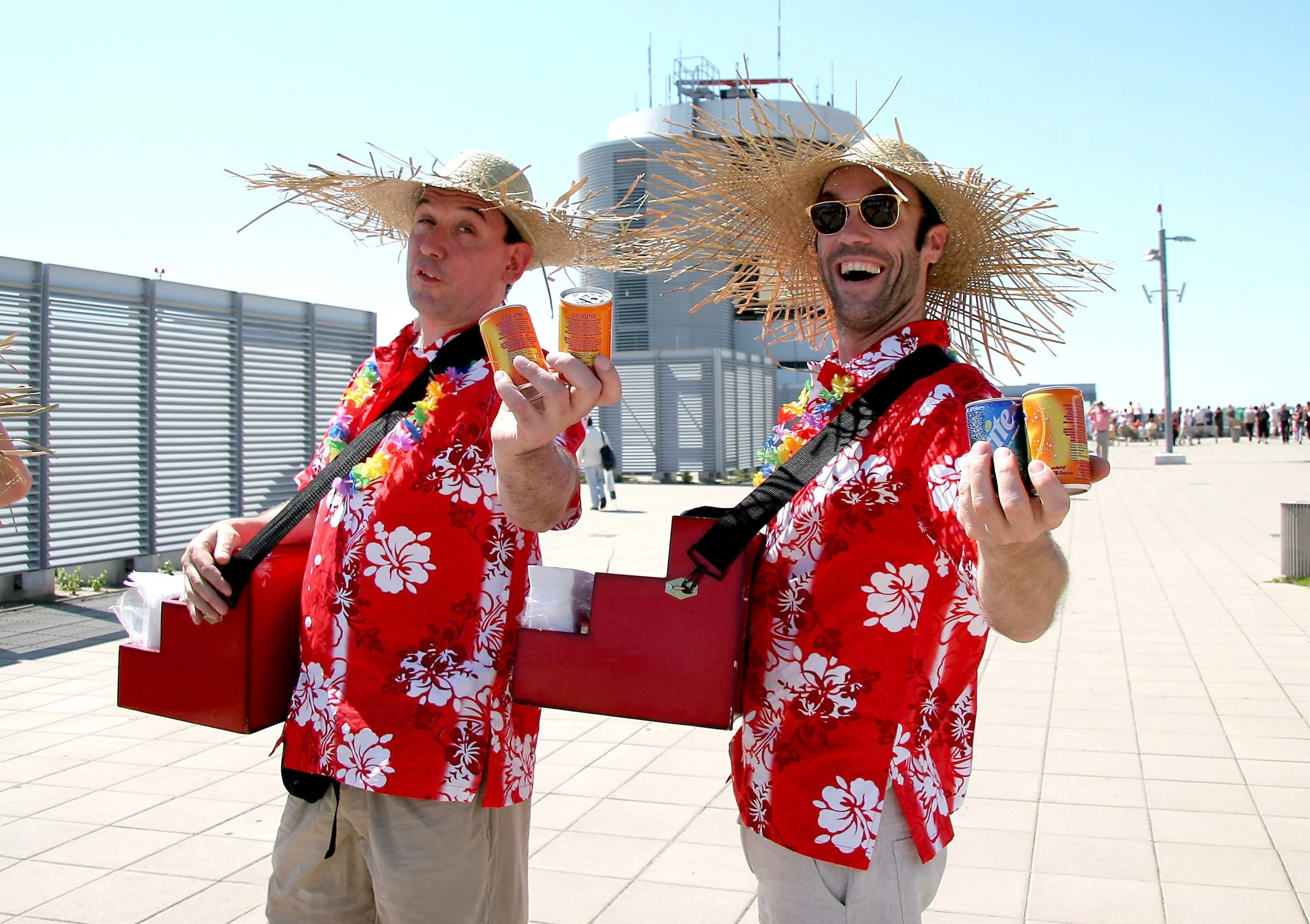 Zwei Männer in Hawaii-Hemden mit Hibiskusmuster, großen Strohhüten, Sonnenbrillen und Lei-Ketten, die auf einer rooftop-Terrasse mit Blick auf die Stadt stehen, hält jeder eine Dose Erfrischungsgetränk in der Hand.