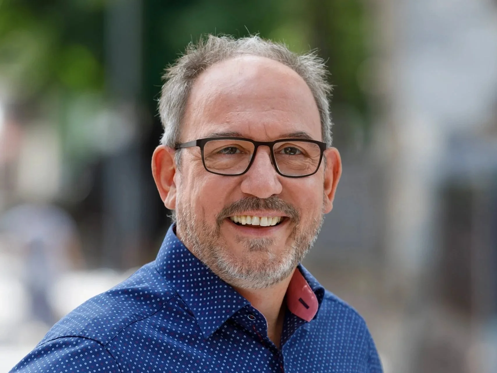 Lächelnder Mann mit Brille und grauem Bart, trägt ein blautes Hemd, auf einer belebten Straße im Freien