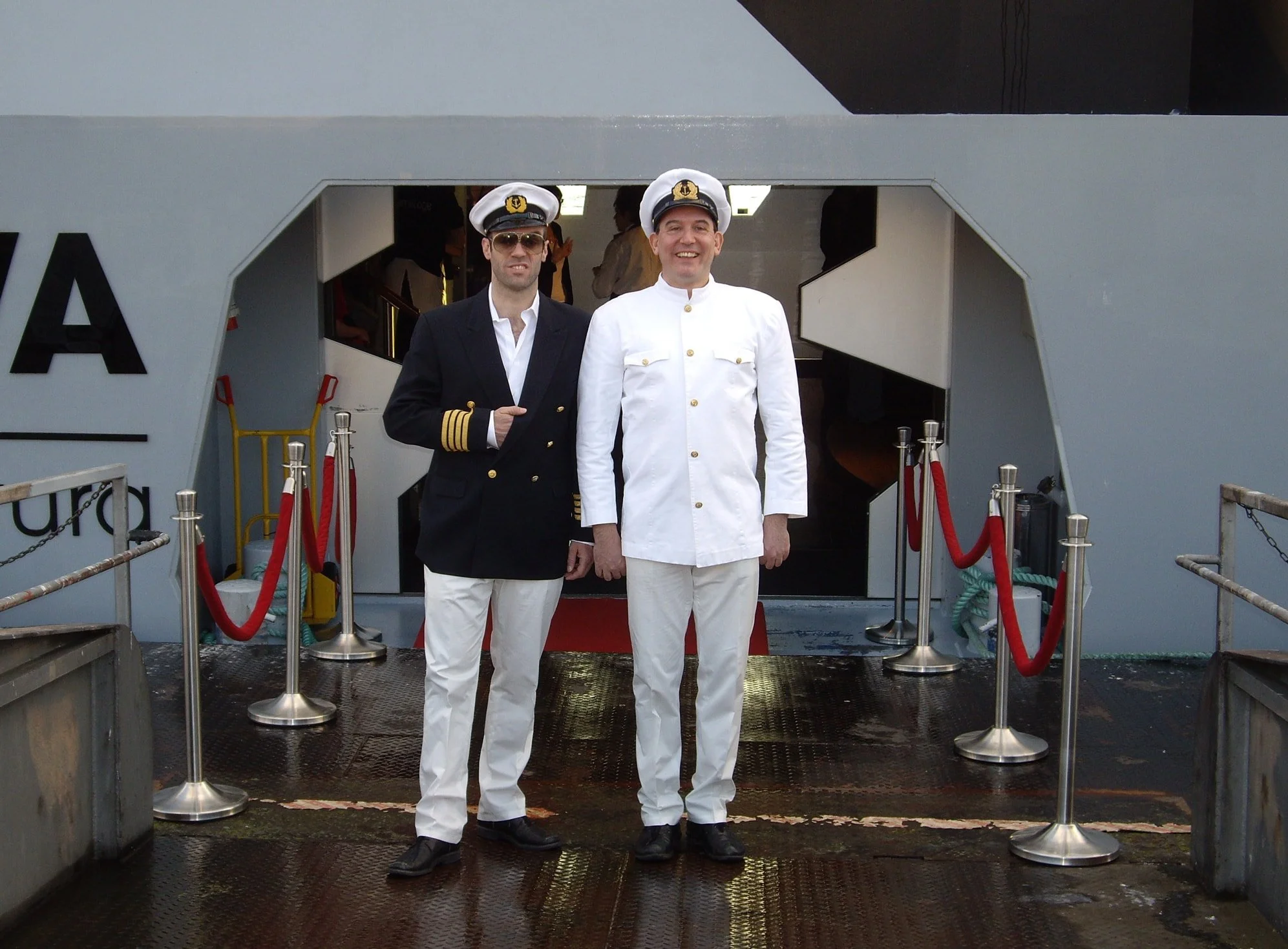 Zwei Männer in Uniformen stehen vor einem Kreuzfahrtschiff, das als Besatzungsmitglieder gekleidet sind. Eines trägt einen schwarzen Anzug mit Schulter- und Armschildern, das andere ein weißes Marineuniform. Sie stehen auf einem Metallsteg mit einem roten Teppich und Absperrungen.