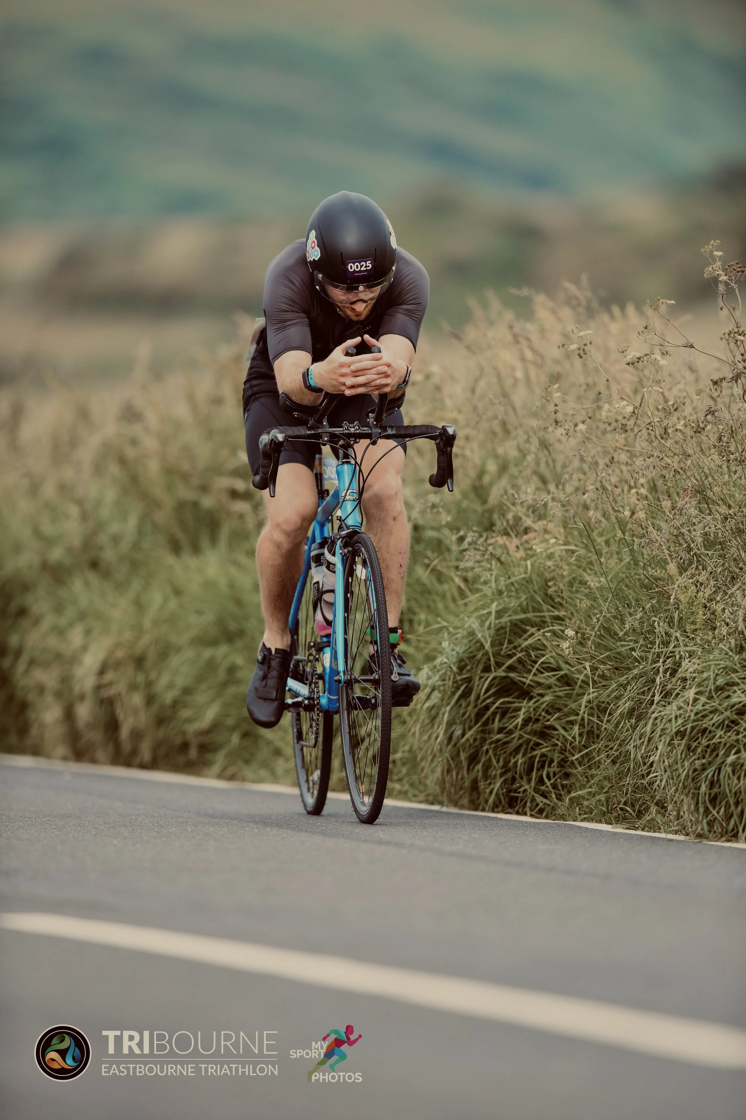 A man in athletic gear riding a blue road bike on a paved path surrounded by tall grass. He is wearing a black helmet and black clothing, and appears to be looking down at his phone while cycling.