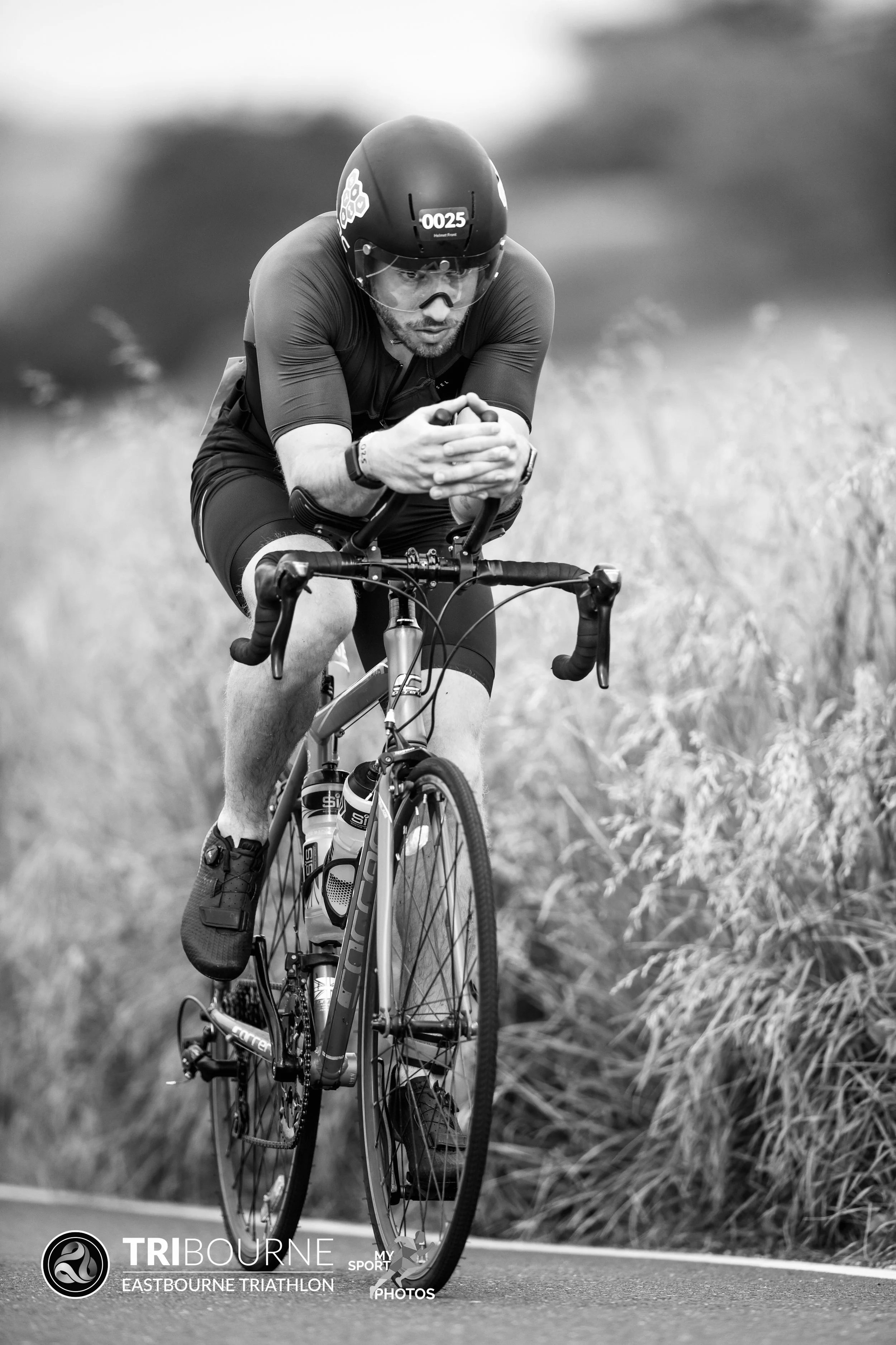A man in athletic gear rides a bike on a road during a triathlon, with tall grass on either side.