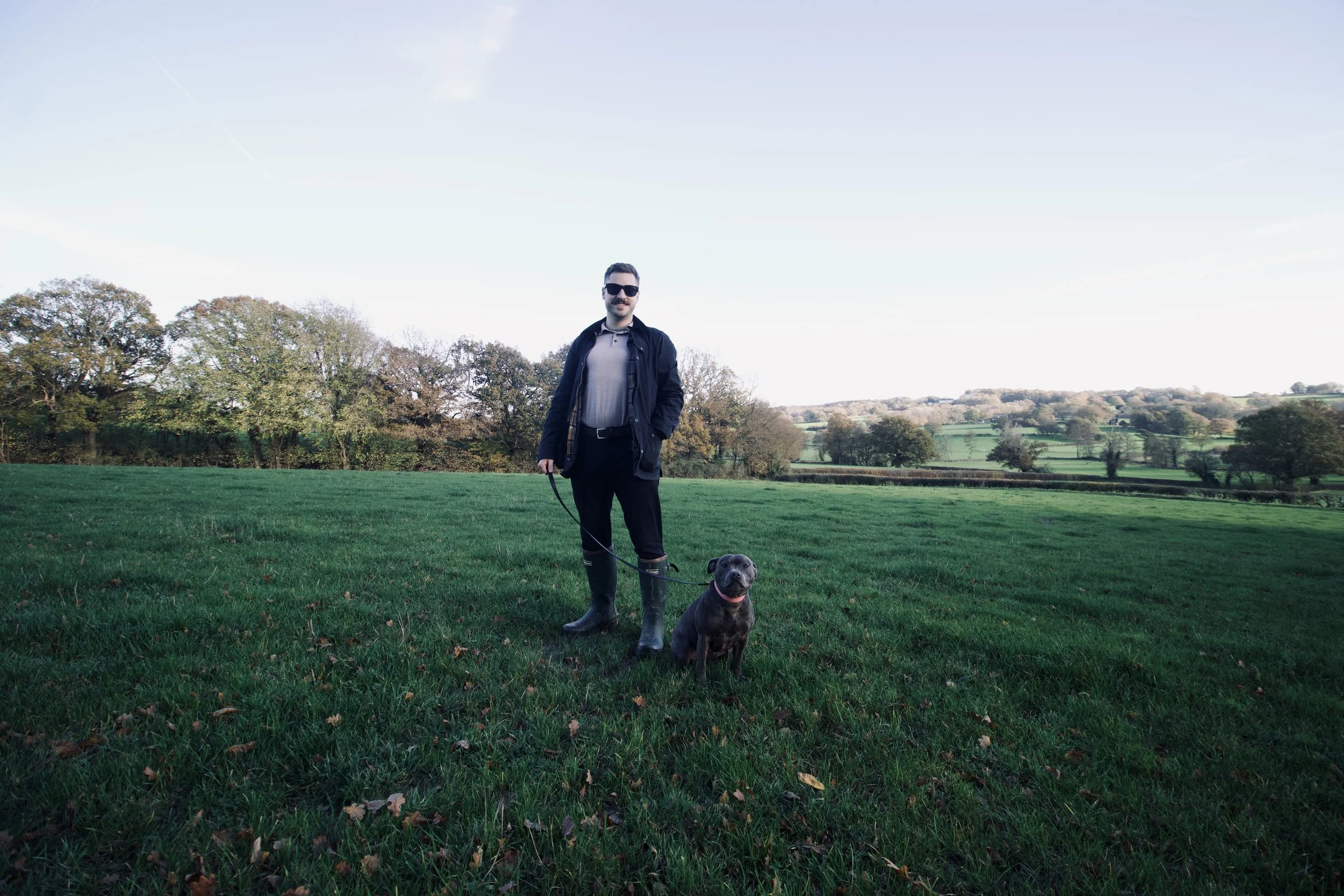 A man with sunglasses standing on a grassy field, holding a leash attached to a black dog, with trees and rolling hills in the background under a clear sky.
