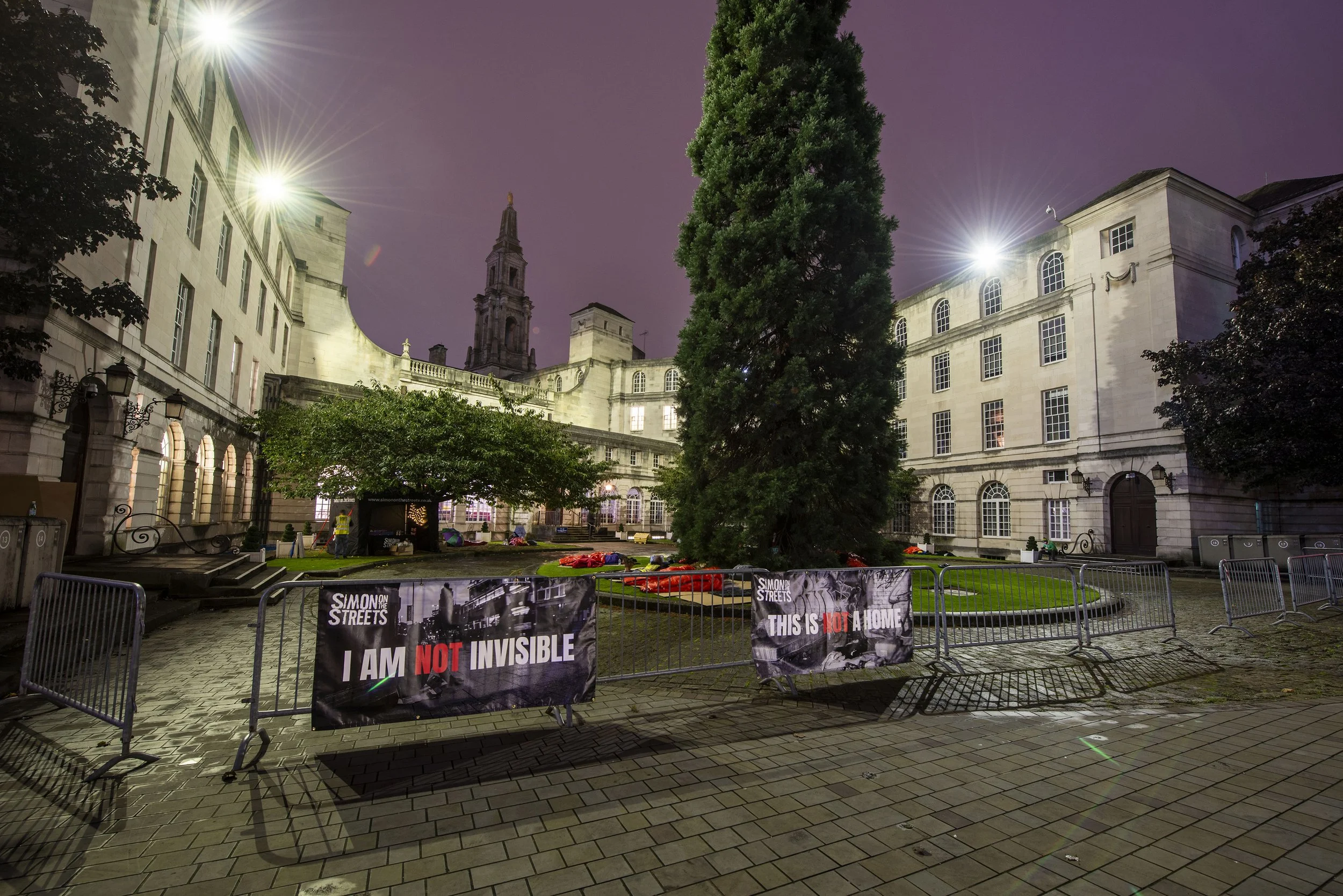 Nighttime scene of a historic building with a tall steeple, illuminated by bright lights, with barriers and protest signs in the foreground. The signs read 'I am NOT invisible' and 'This is NOT a home.'