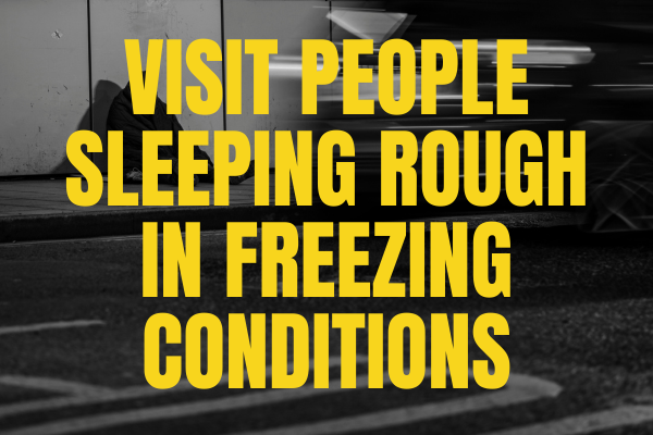 A sign with bold yellow text that reads: "Visit people sleeping rough in freezing conditions," placed in front of a street scene with parked cars.