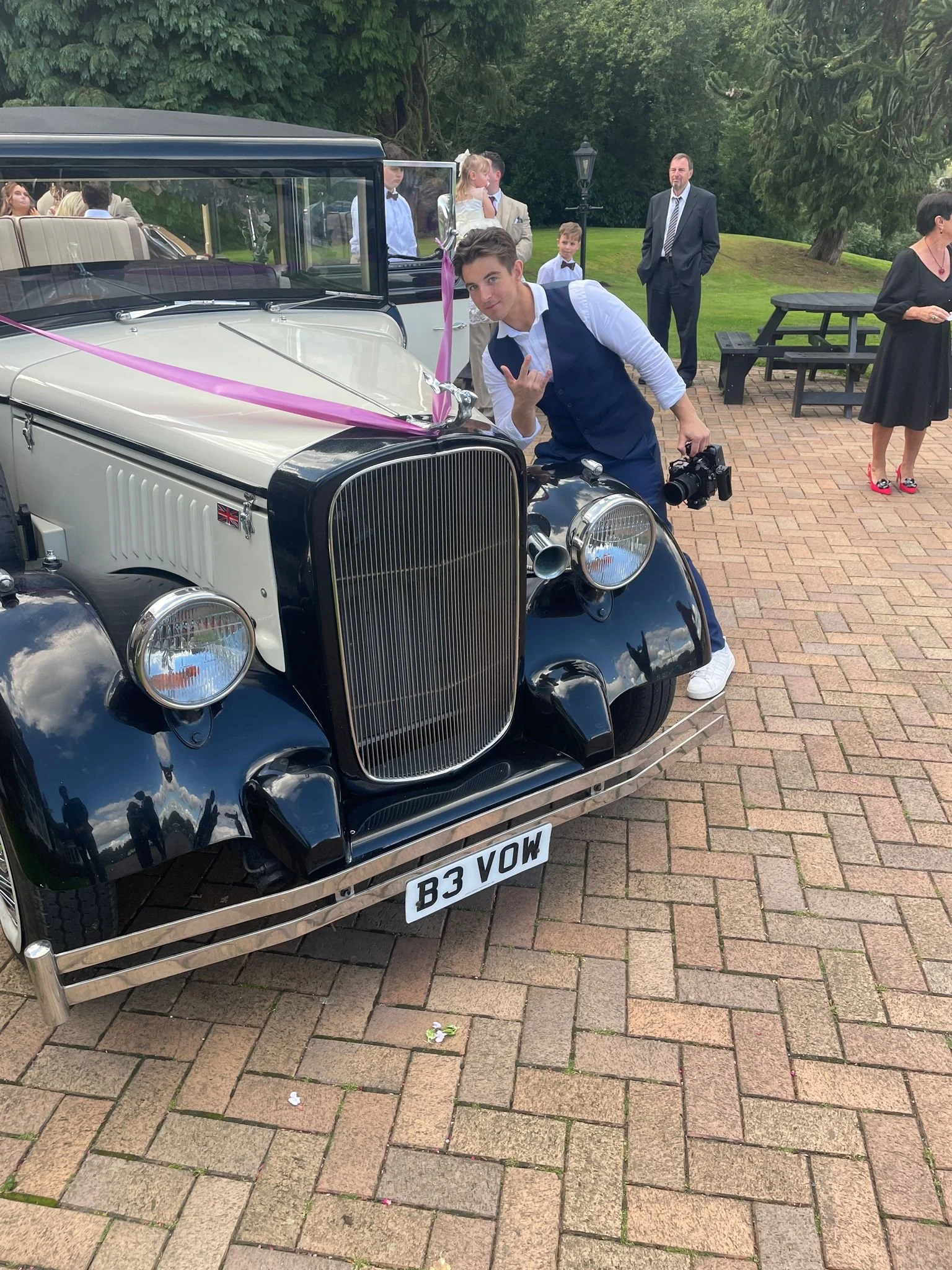 A young man in a white shirt and navy vest leans on the front of a vintage black and cream car decorated with pink ribbons, at what appears to be a wedding or special event, with other people in formal attire in the background.