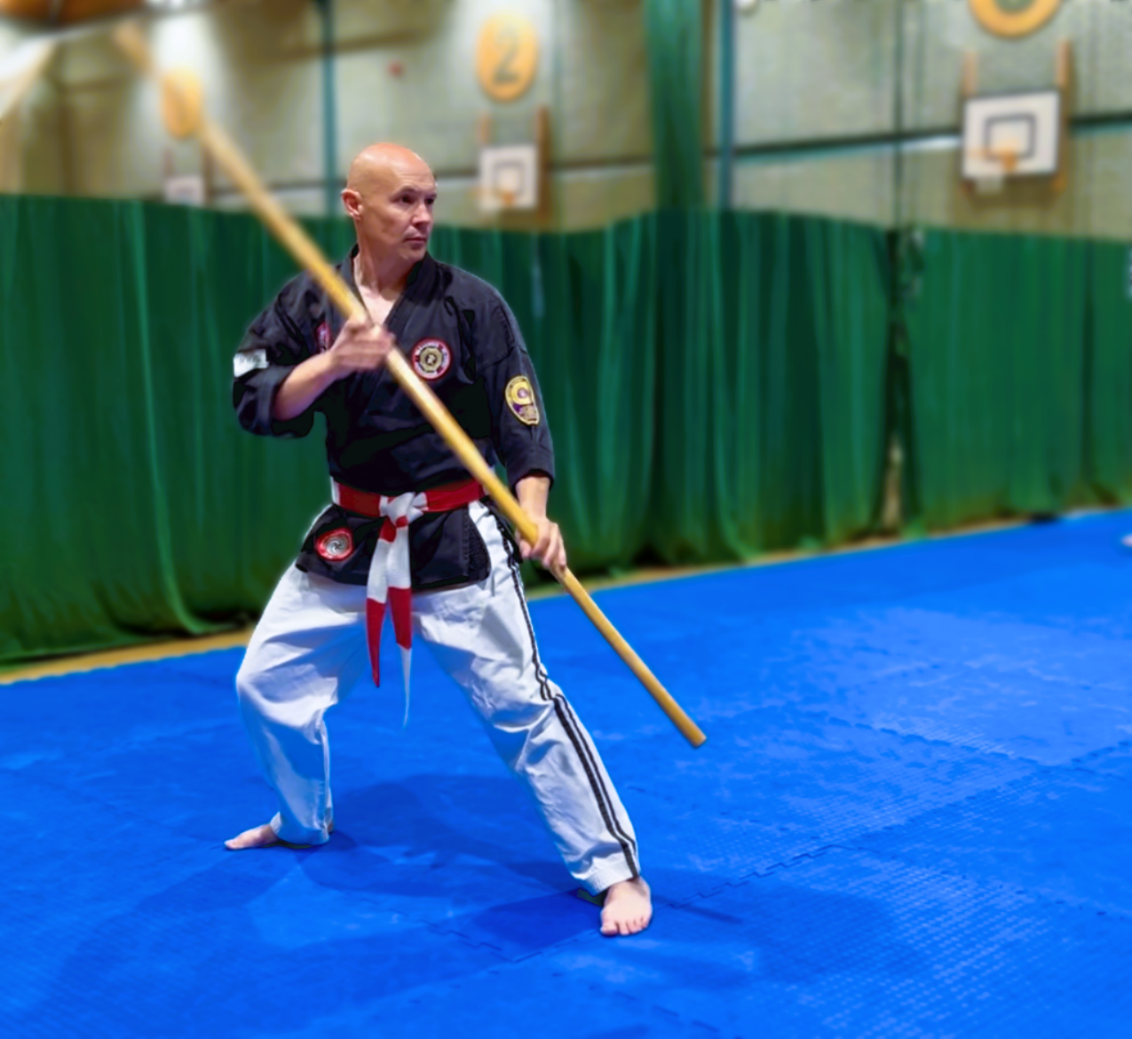 A martial artist practicing with a bo staff in a gym, wearing a black uniform with patches and a red and white belt, on a blue mat.