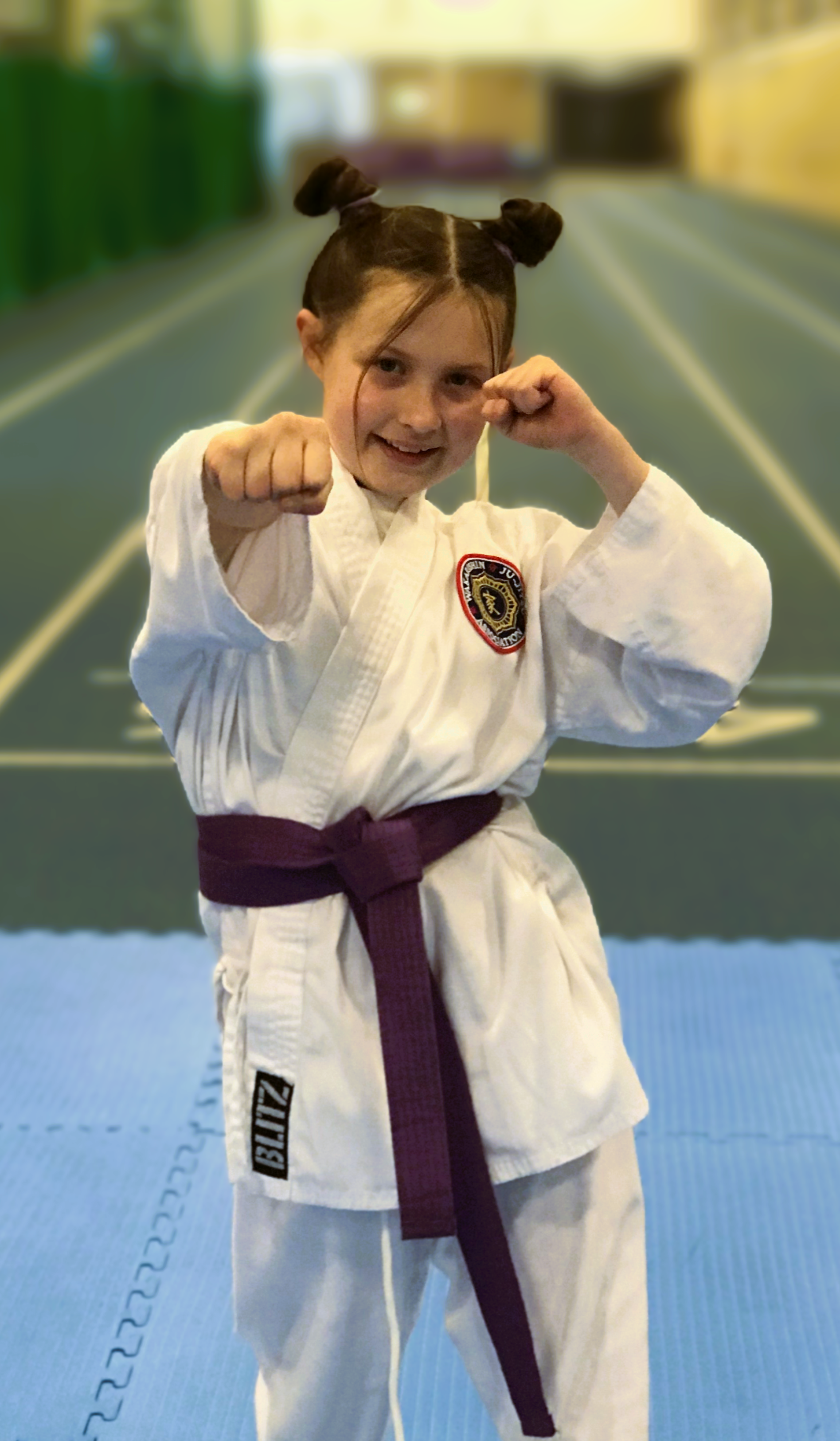 Young girl in a martial arts uniform with a purple belt, making a fighting pose in a dojo or training area.