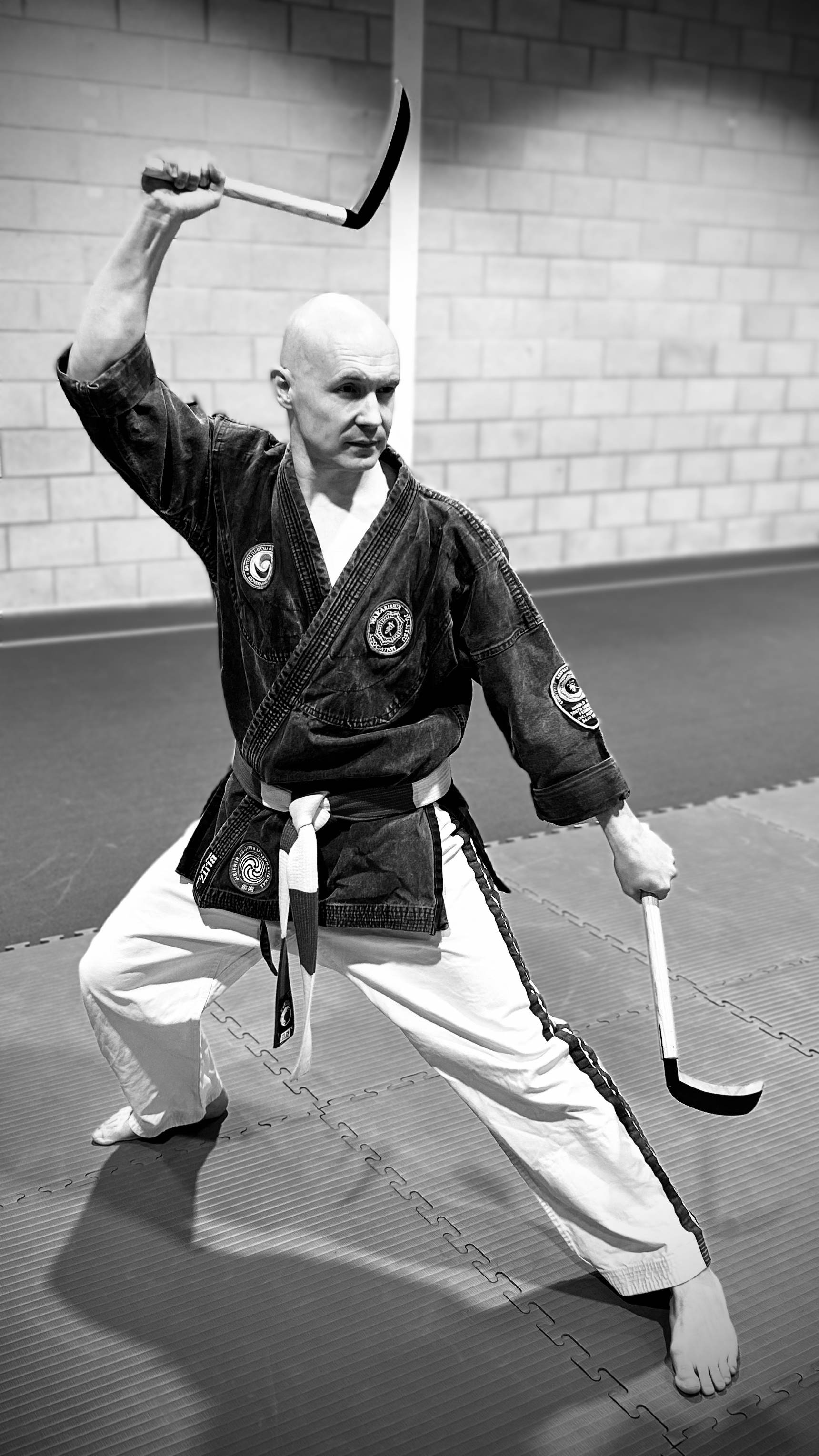 A martial artist in a black gi with patches, white pants, and a black belt demonstrating a martial arts stance while holding two sticks, in a dojo with a brick wall background.