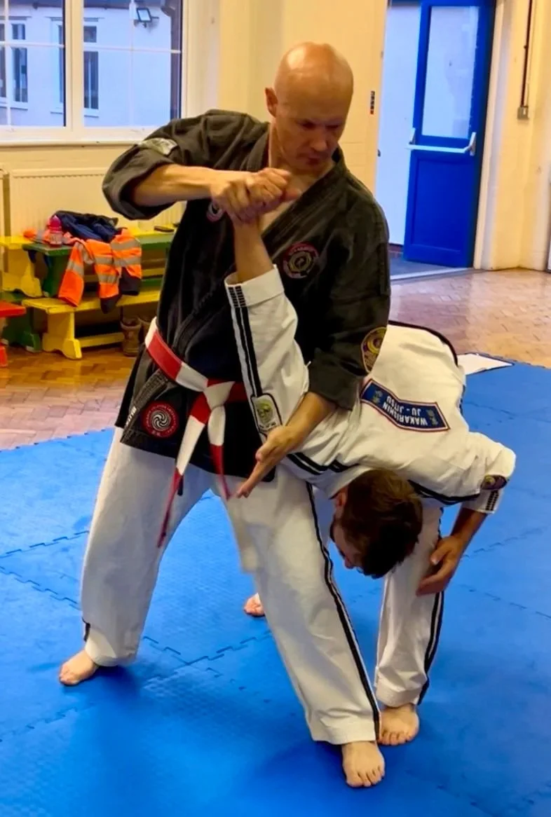 Two martial artists practicing Jiu-Jitsu, with one in a black gi and the other in a white gi, on a blue mat in a training room.