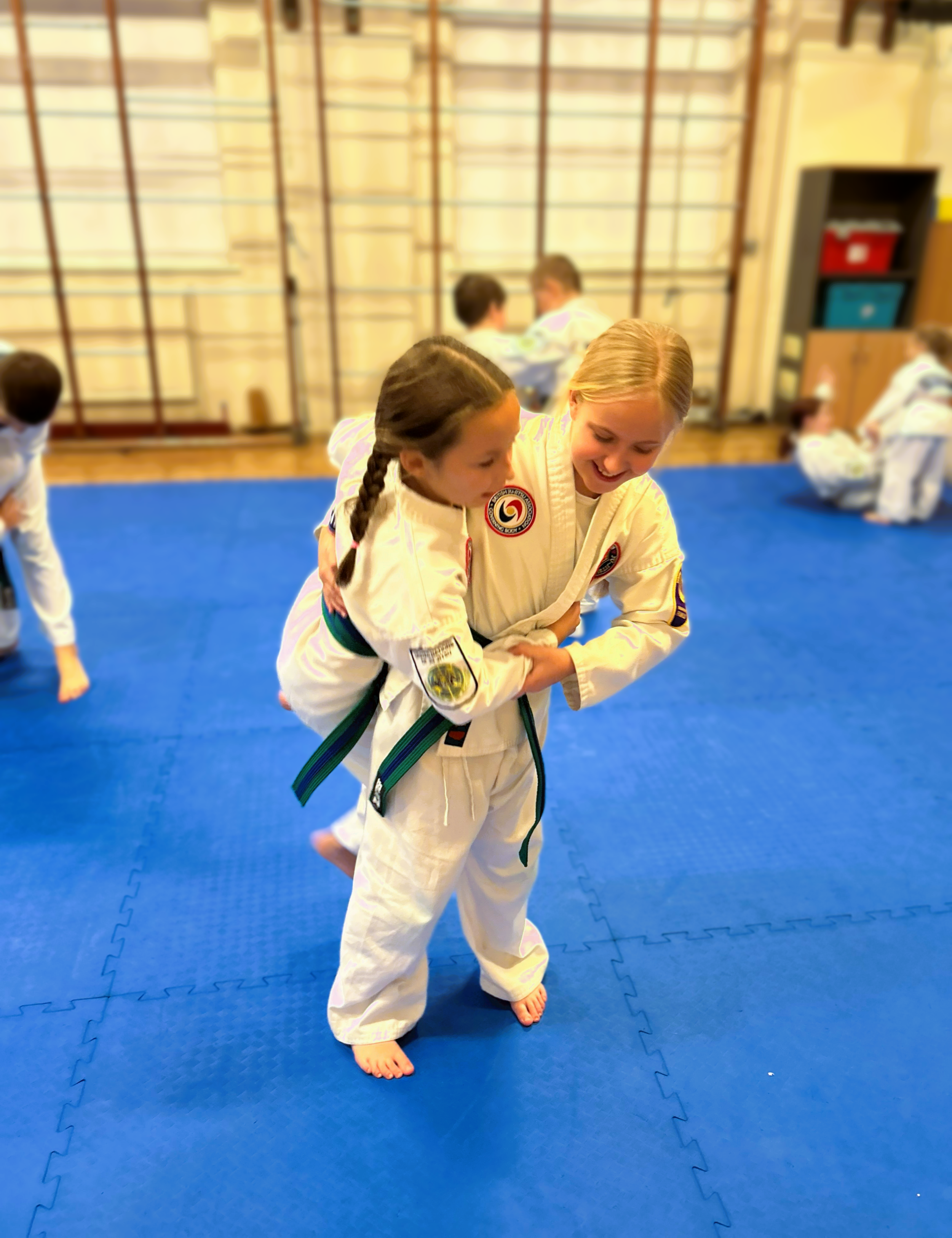Two young girls practicing Japanese Ju-Jitsu in a martial arts dojo, one girl is helping the other learn a grappling technique, with other children practicing in the background.