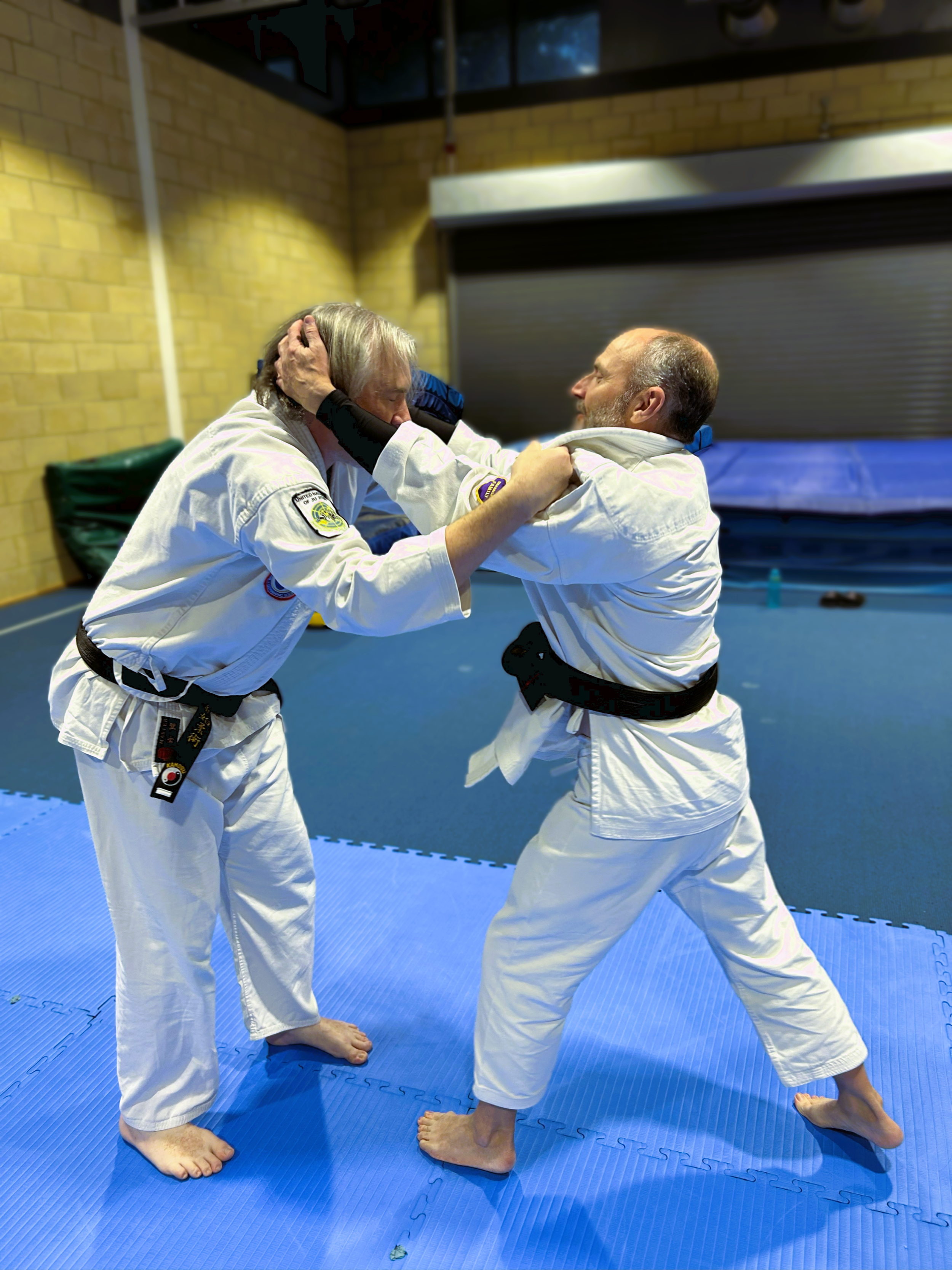 Two men practicing martial arts in a dojo, wearing white uniforms and black belts, engaging in a sparring drill on blue mats.