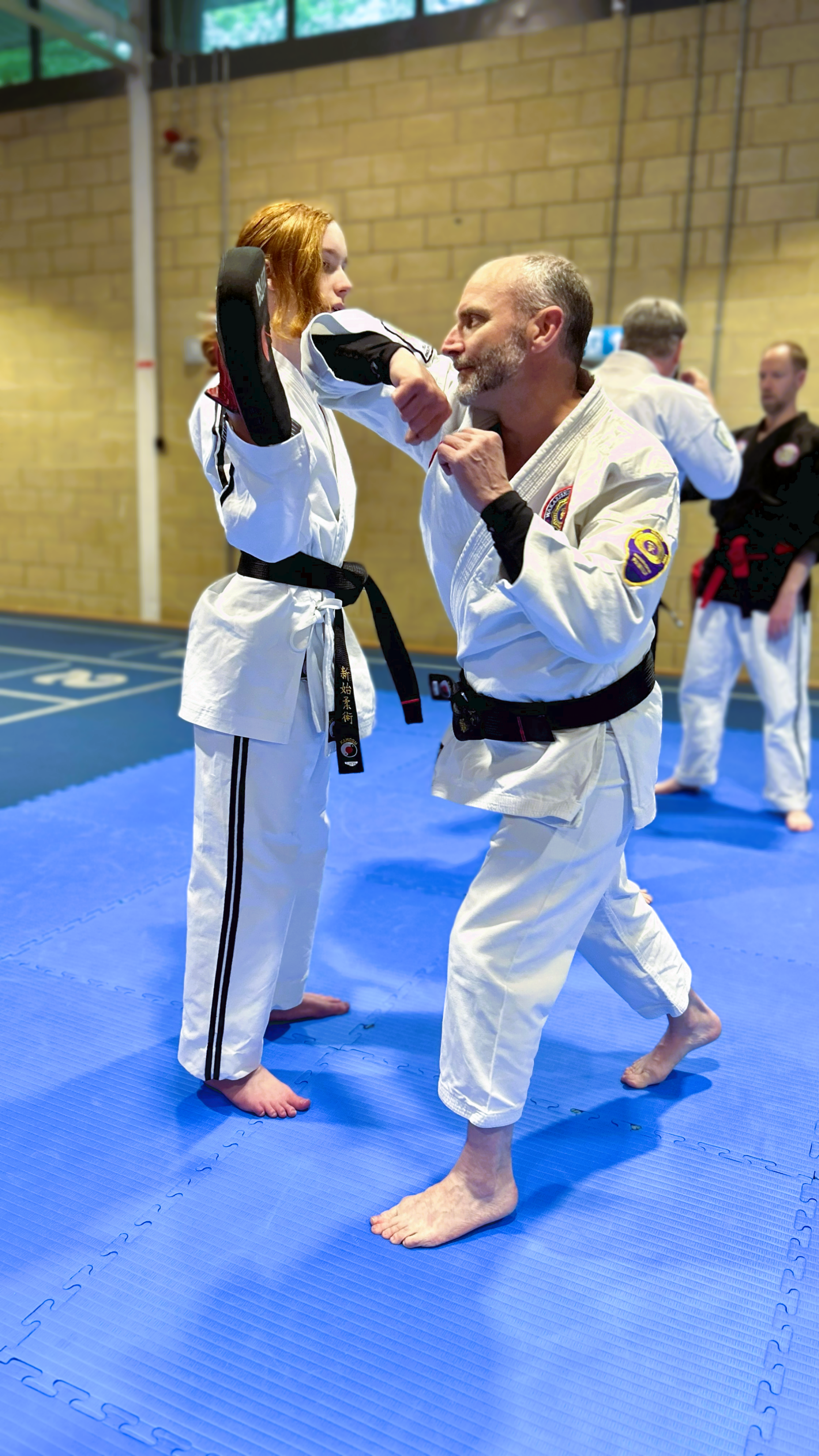 Martial arts students practicing on blue mat, one man performing an elbow while an instructor blocks with a pad, all wearing traditional martial arts uniforms.