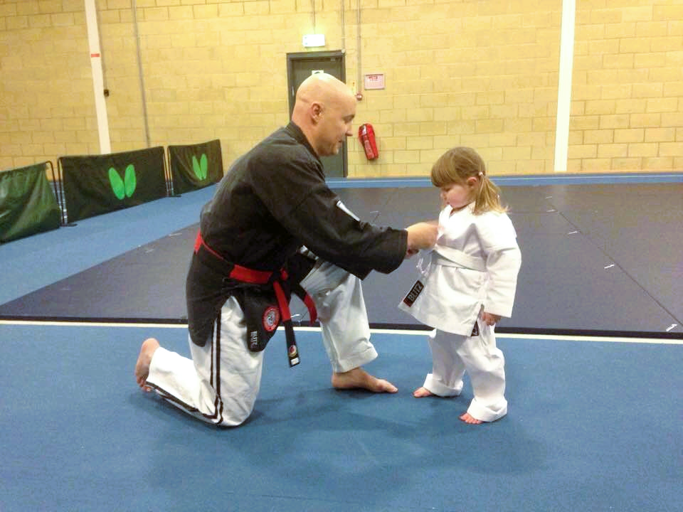 An adult and a young girl practicing martial arts on a blue mat in a gym, with the adult kneeling and the girl standing, both in martial arts uniforms.
