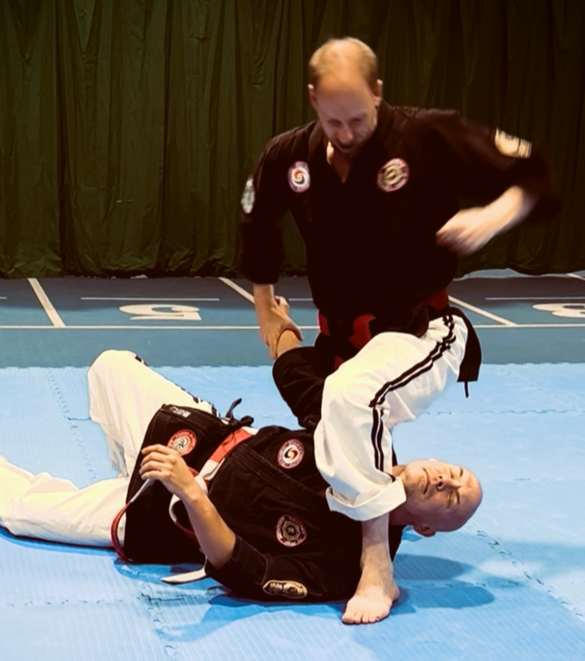 Two martial artists practicing Japanese Ju-Jitsu on a blue mat; one standing and applying a choke to the other lying on the mat, both wearing black gis with patches.