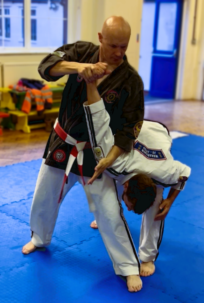 Two martial artists practicing Jiu-Jitsu, with one in a black gi and the other in a white gi, on a blue mat in a training room.