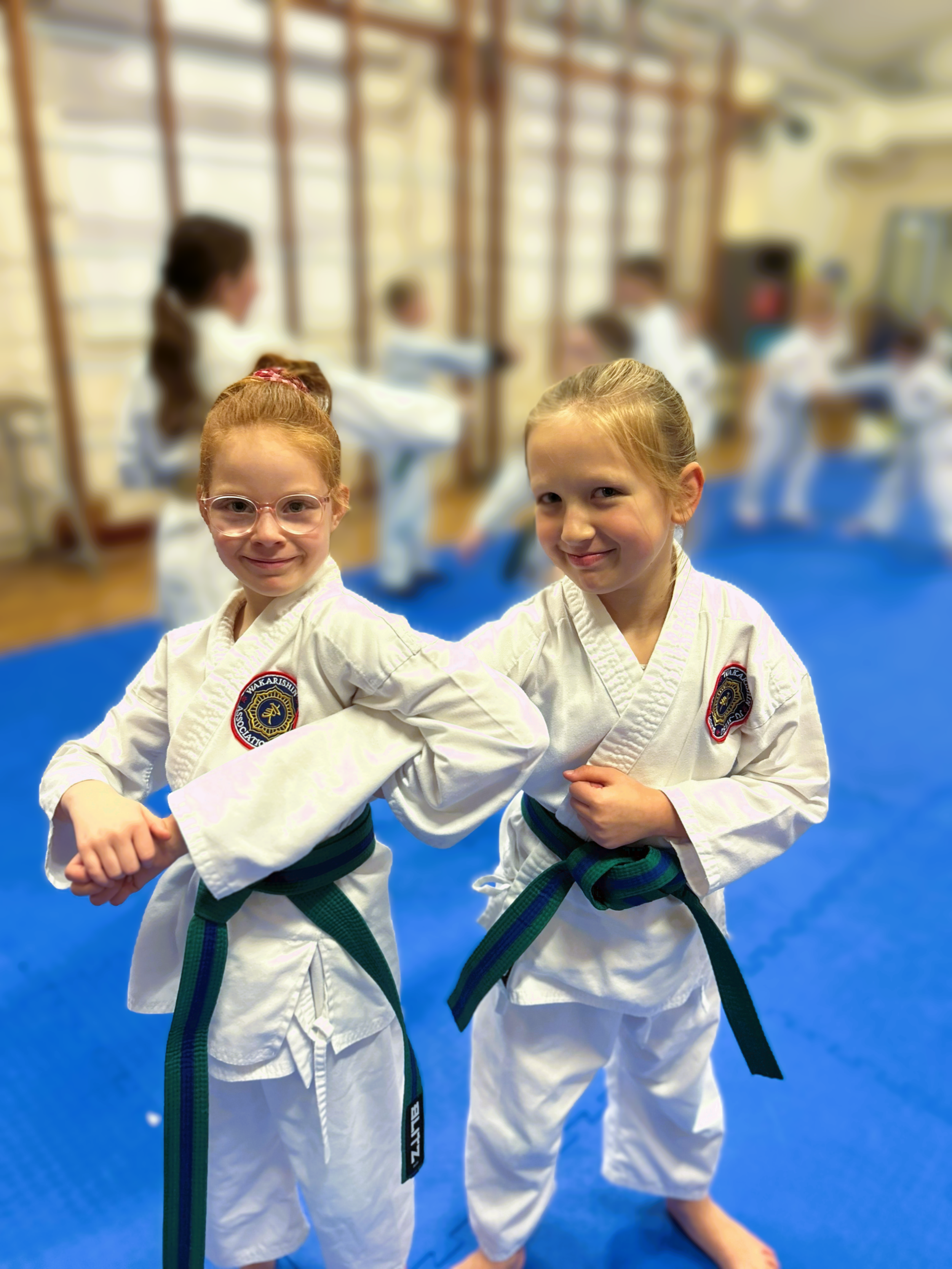 Two young girls in martial arts uniforms with green belts, smiling and posing in a dojo with other children practicing martial arts in the background.