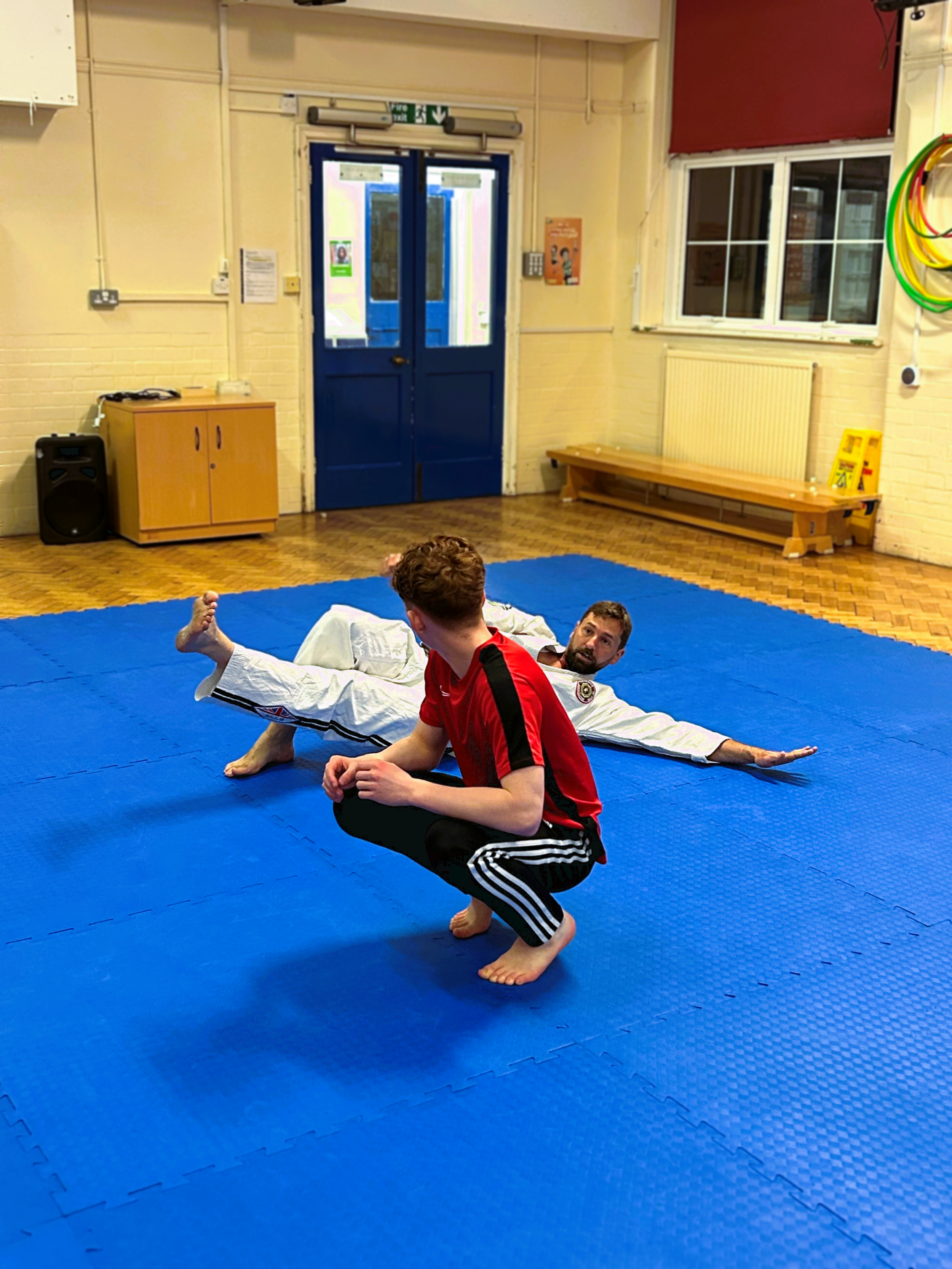 Two men practicing martial arts on a blue padded floor in a gym, with one in a white gi lying on the floor and the other in a red shirt and black pants squatting.