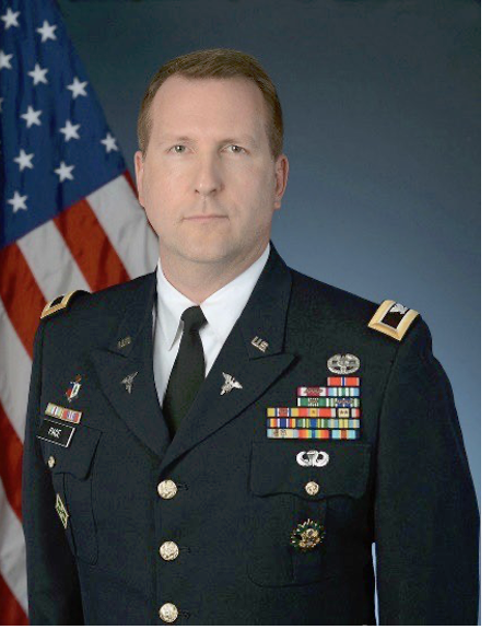 A man in a U.S. military dress uniform standing in front of an American flag.