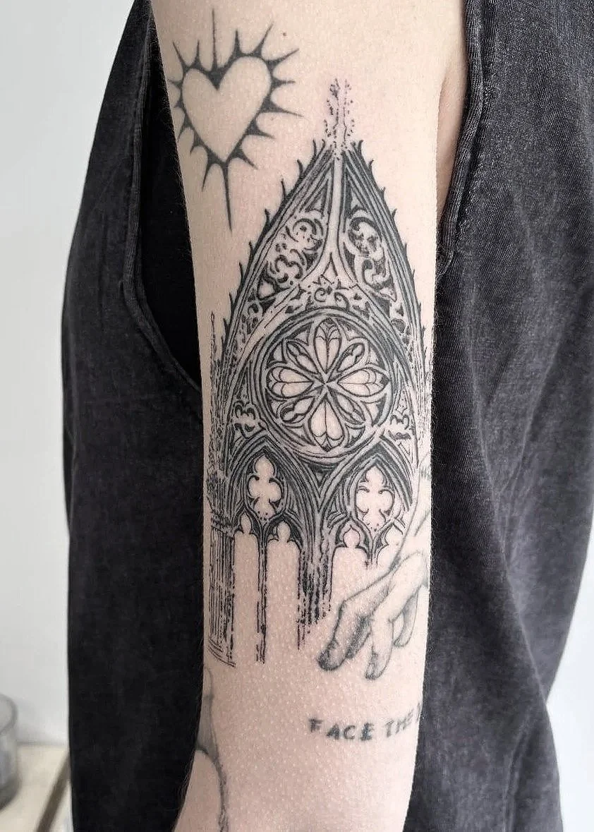 Tatuaje de una figura arquitectónica gótica, una mano apuntando, un corazón con rayos y parte del texto que dice "FACE THE".