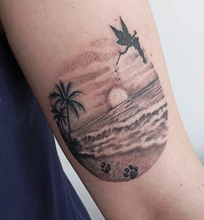 Tatuaje en el brazo que muestra una escena de playa con palmeras, olas, el sol y un dragón volando en el cielo.