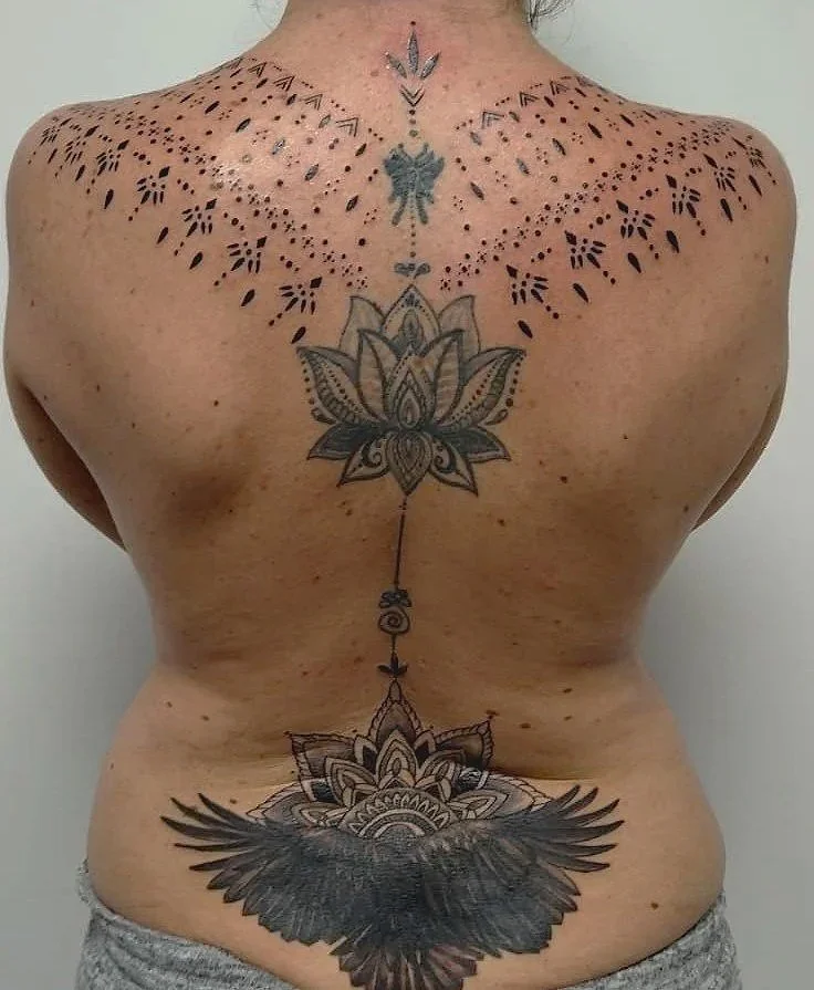 Tatuaje de un personaje con alas de plumas negras, flanqueado por patrones decorativos en la espalda y en la parte baja de la espalda, con un diseño de flor de loto en el centro de la espalda.