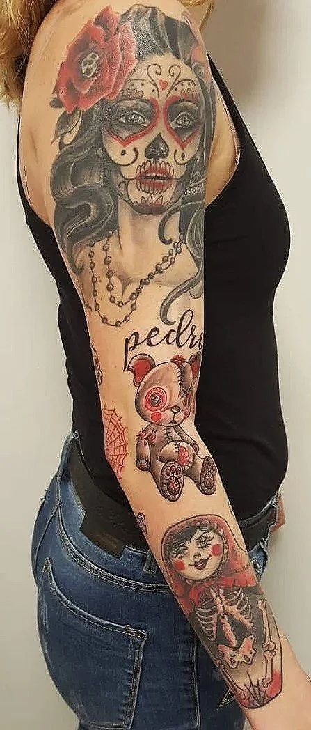 Tatuajes en el brazo de una persona que incluyen una mujer con maquillaje estilo calavera mexicana, una cabeza de muñeco, una niña con ojos en espiral, una calavera con pelo rizado y una telaraña, todo en estilo de arte tradicional con colores vivos 