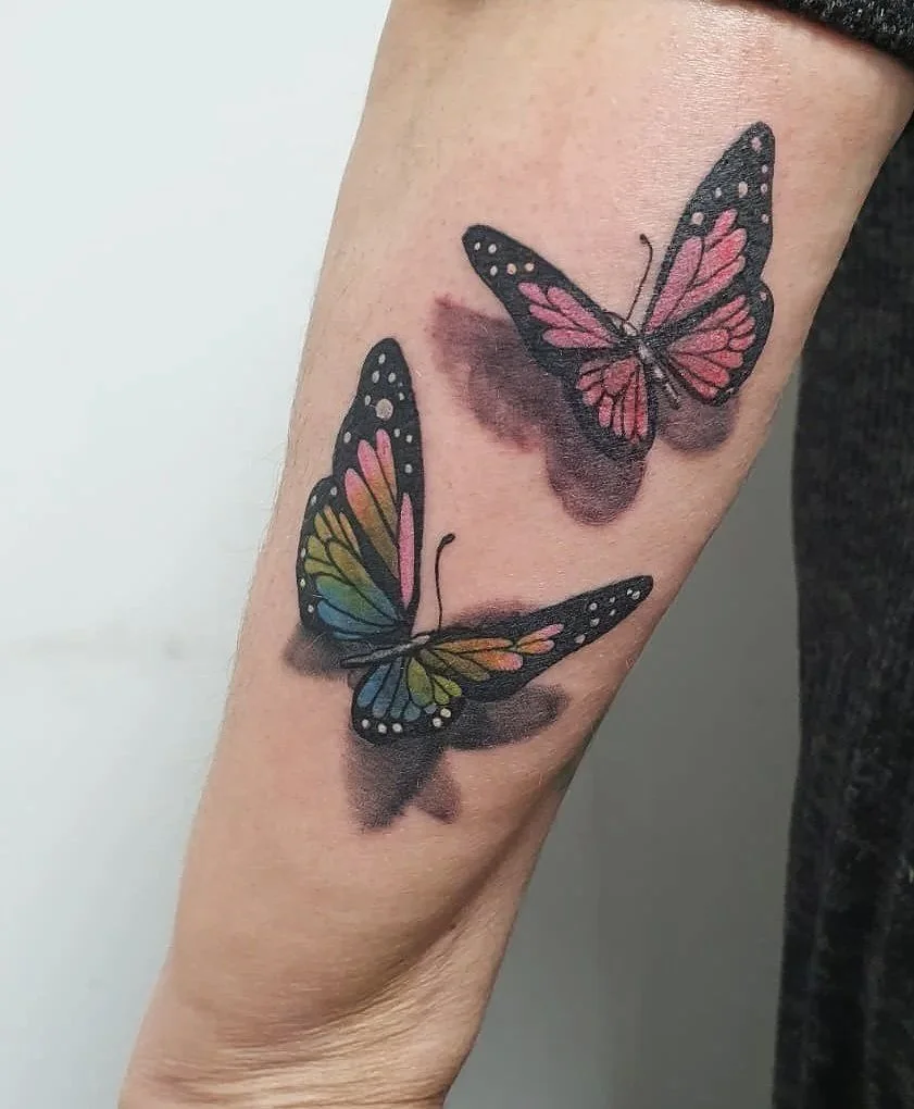 Tatuaje de dos mariposas en el antebrazo, con alas coloridas en tonos rosa, azul, verde y negro, y sombras debajo para dar efecto de profundidad.